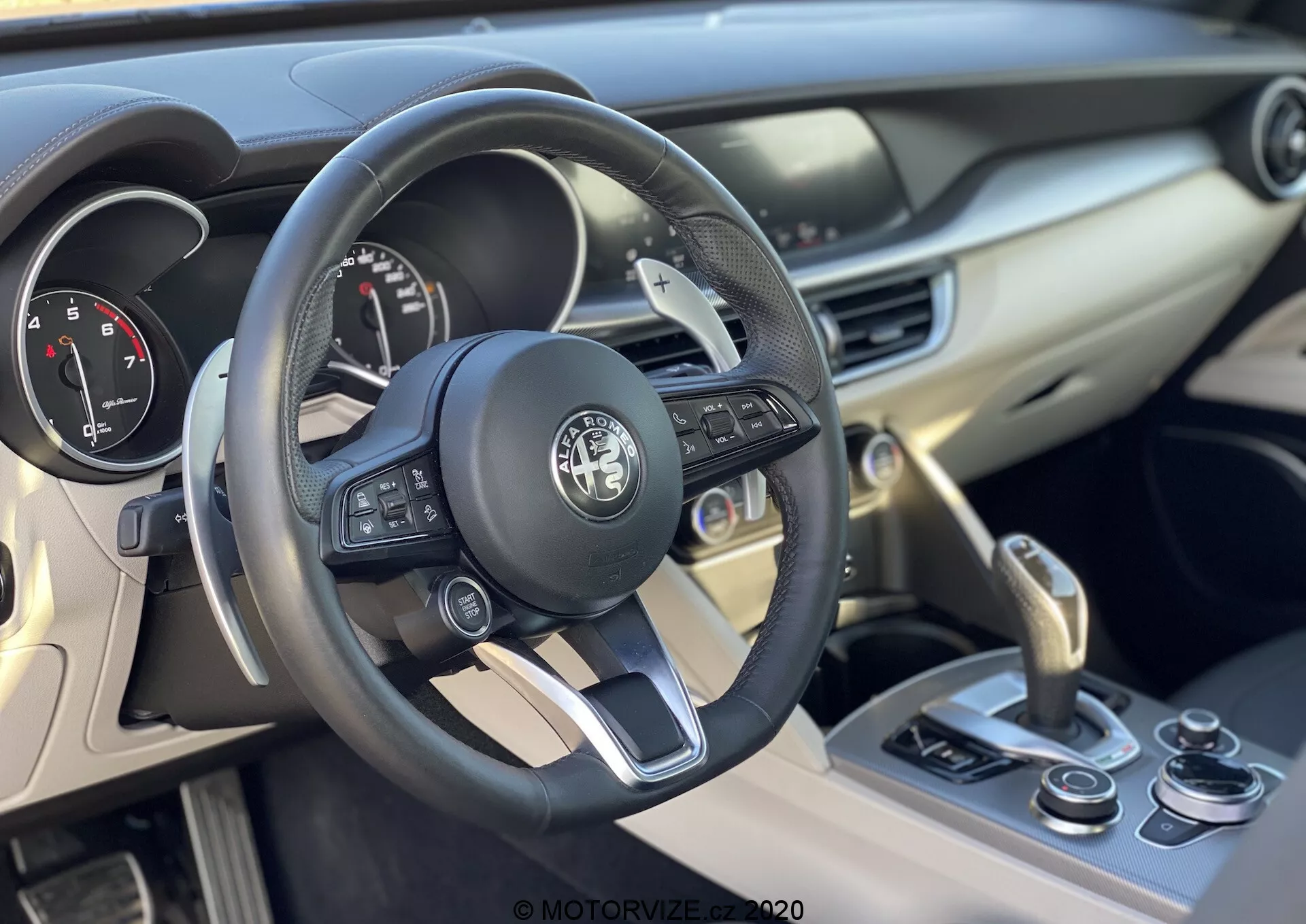 2017-es Alfa Romeo Stelvio belső nézete, a kormánykerékre és a műszerfalra fókuszálva. A bőrkormány többfunkciós gombokkal van ellátva a multimédiához, hívásokhoz és sebességtartó automatához, és központi helyen található a gyártó emblémája. A kormánykerék mögötti műszerfal analóg óraja tartalmazza a sebességmérőt és a fordulatszámmérőt, amely valószínűleg egy kis digitális kijelzővel is rendelkezik további információk megjelenítéséhez. A jobb oldalon található középkonzol légbeömlő nyílásokat, egy motorindító-stop gombot és egy sebességváltókart tartalmaz, amely automata sebességváltót jelez. Az egyéb vezérlők, mint például a vezetési mód kiválasztó, is megtalálhatók itt. A belső tér anyagai között megtalálhatók a puha tapintású műanyagok és a bőr, fémes díszítésekkel együtt, amely a vezető ergonómiájára és kényelmére helyezi a hangsúlyt.