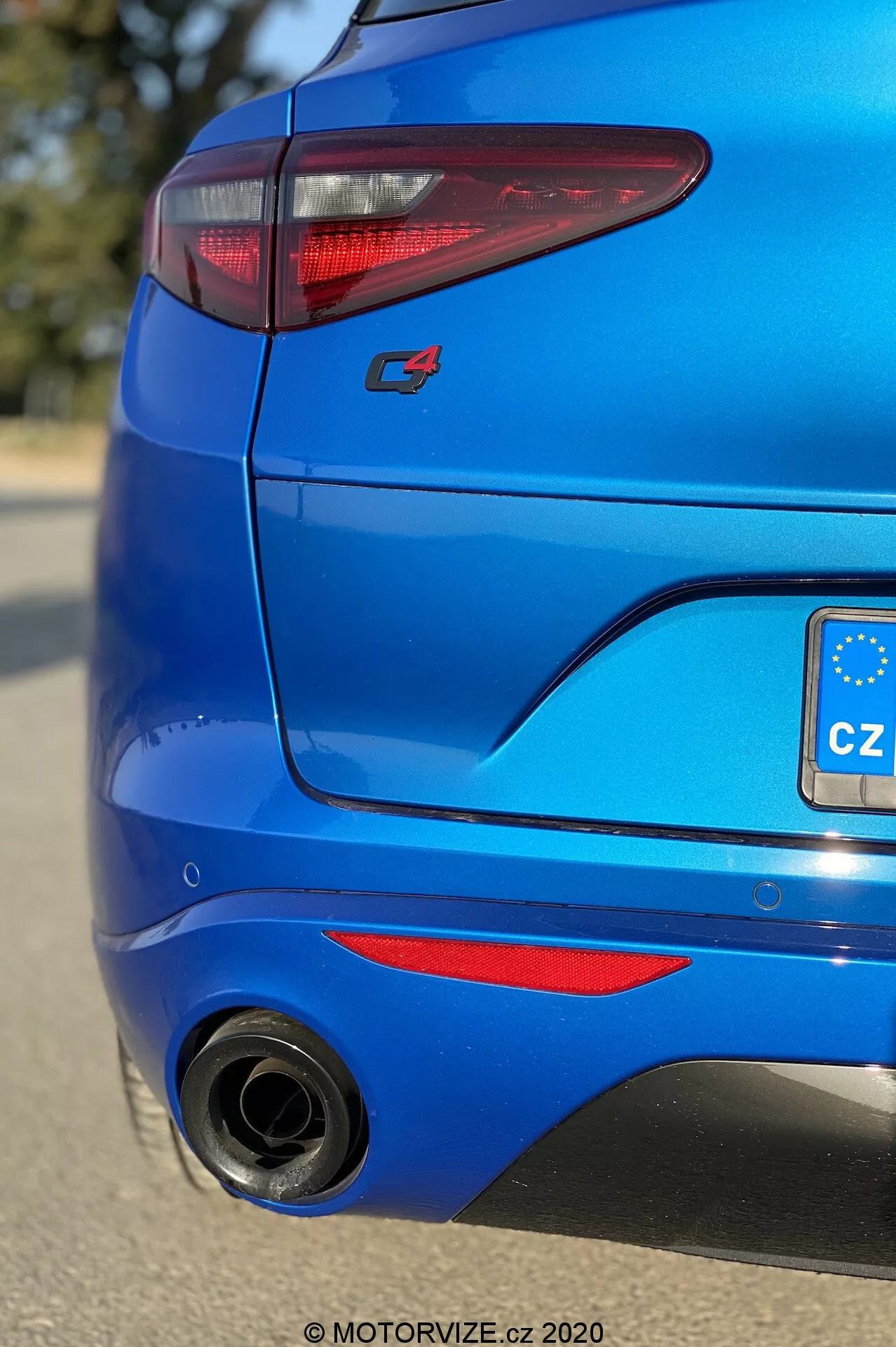 Közelkép egy élénkkék 2017-es Alfa Romeo Stelvio hátulsó részéről, alacsony szögből, kissé jobbra fókuszálva a hátsó sarokra. Jellemzői közé tartoznak az elegáns C-alakú LED hátsó lámpák, egy sportos piros 'S' badge, egy diffúzor-szerű elem fekete betéttel az hátsó lökhárítón, egy nagy kör alakú kipufogócső bal oldalon, fémes kék festés, piros reflektorok és egy kis tetőspoiler, kiemelve a jármű modern kialakítását és sportos esztétikáját.