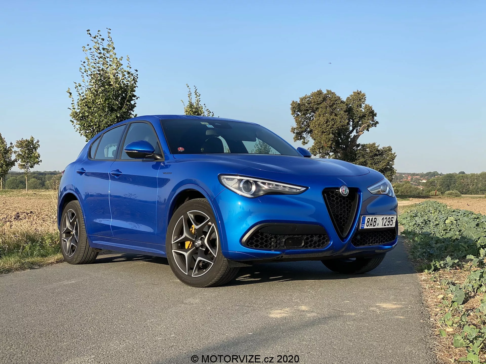 A képen egy kék, 2017-es Alfa Romeo Stelvio sport szedán látható, amely az út szélén parkol egy vidéki vagy félvidéki környezetben. Az autó szögben helyezkedik el, így a háromnegyedes nézetből jól látható az eleje és bal oldala, kiemelve a jellegzetes hűtőrácsot és fényszórókat. Láthatóak a modern, elegáns világítás, a formázott motorháztető dizájnja, a fekete, többlépcsős könnyűfém felnik és a teljesítményorientált féknyergek. A természetes fény azt jelzi, hogy a fotó nappal készült, valószínűleg reggel vagy késő délután, fák és nyílt mezők láthatók a háttérben, nyugodt hangulatot teremtve.