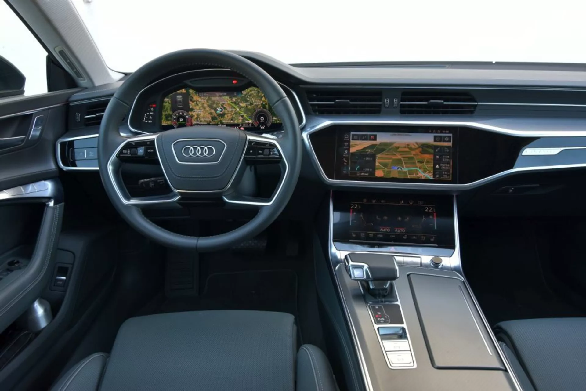 2018-as Audi A7 Sportback belső nézete a vezető műszerfalára és középkonzol területére összpontosítva, bemutatva egy háromküllős multifunkciós kormányt Audi logóval, digitális Virtual Cockpit műszerfalat navigáció megjelenítéssel, két kijelzős infotainment rendszert klíma beállításokkal, ergonomikus váltókart és prémium anyagokat, mint a kefélt fém és bőr.
