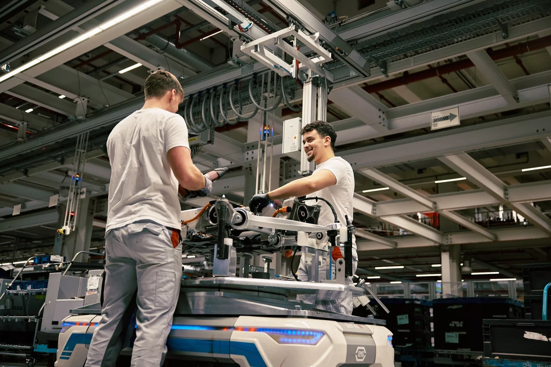 Ipari környezet, ahol két munkás összeszerel vagy karbantart mechanikai berendezéseket egy gyári padlón, mesterséges világítás mellett, magasból vett nézőponttal, amely hangsúlyozza a gyártási környezetet