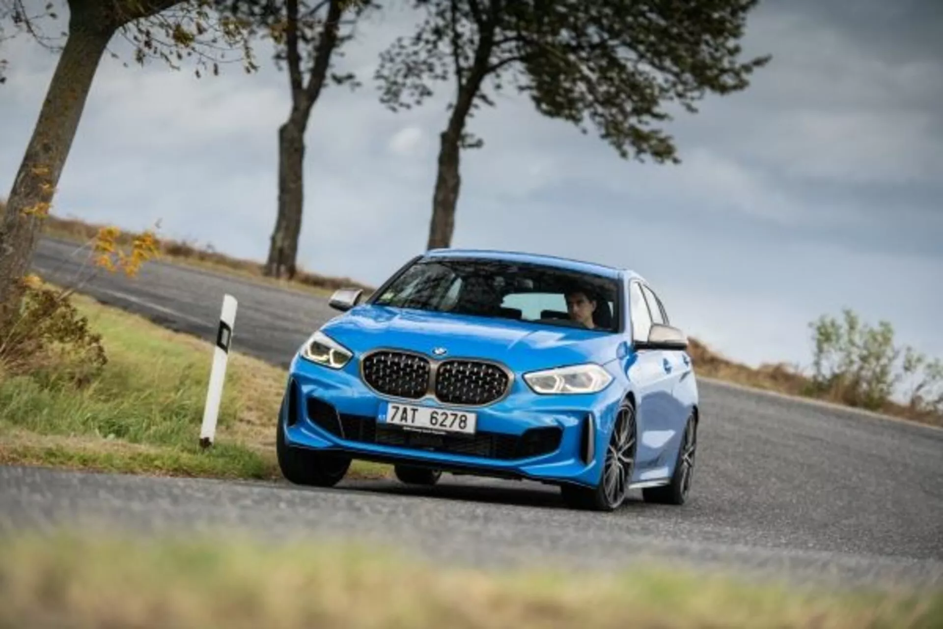 2019-es kék BMW 1-es sorozat M Performance sport szedán alacsony és oldalról nézve ferde perspektívából megörökítve, amely kiemeli agresszív előlapját ikonikus vesemedence rácsokkal, elegáns LED fényszórókkal, nagy légbeömlőkkel a teljesítmény hűtéséhez, alacsony sport felfüggesztéssel, éles kanyarban történő mozgás közben bemutatva a dinamikus kezelhetőséget, aerodinamikus oldalsó tükrökkel, nagy ötvözetű kerekekkel és alacsony profilú abroncsokkal, amelyek kiemelik a sportos státuszt és a tapadási képességeket.