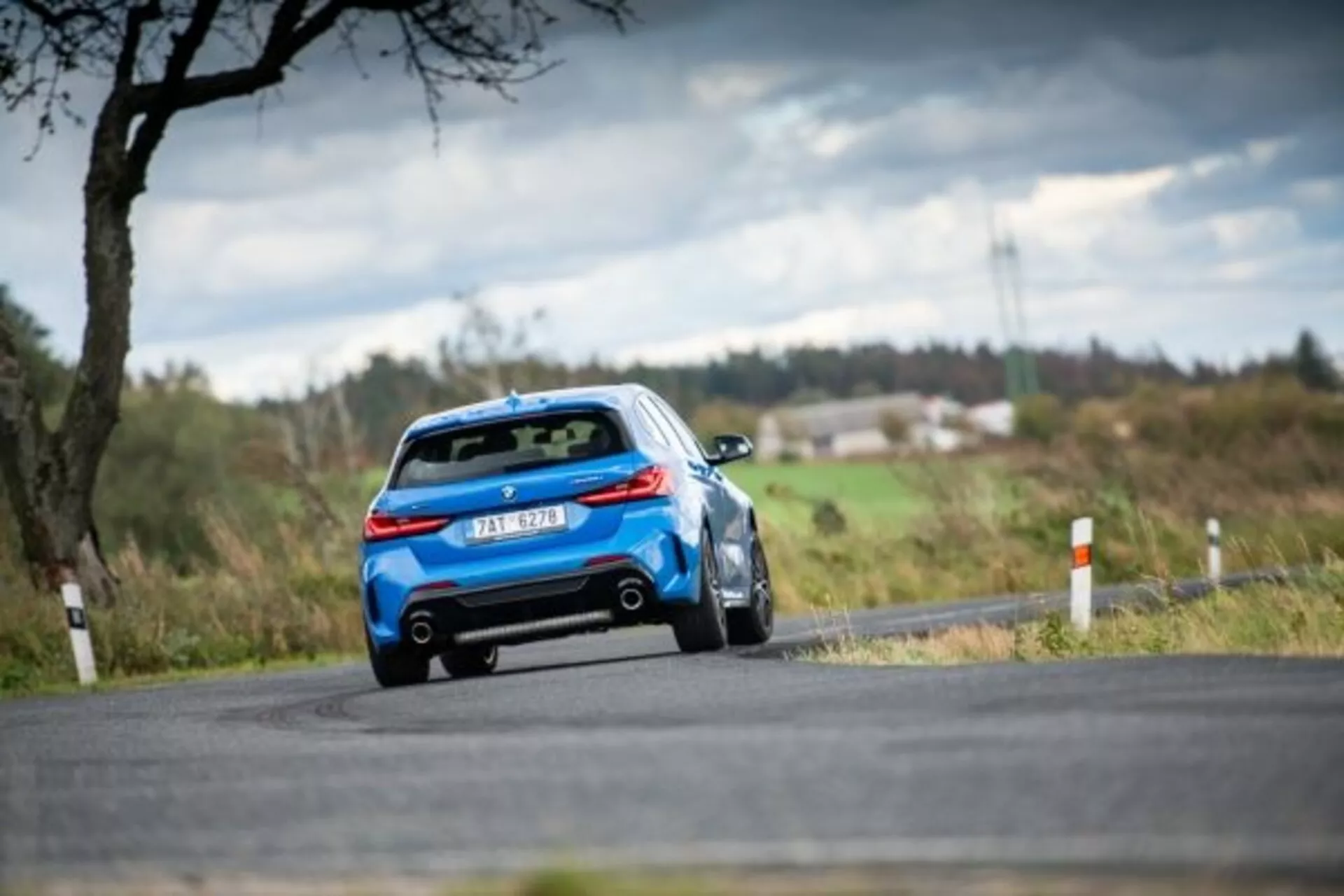 2019-es kék BMW 1-es sorozat M Performance vidéki úton halad, hátsó háromnegyedes nézetben megmutatva az aerodinamikus tervezést hátsó spoilerrel, modern LED hátsó lámpákkal és kettős kipufogórendszerrel