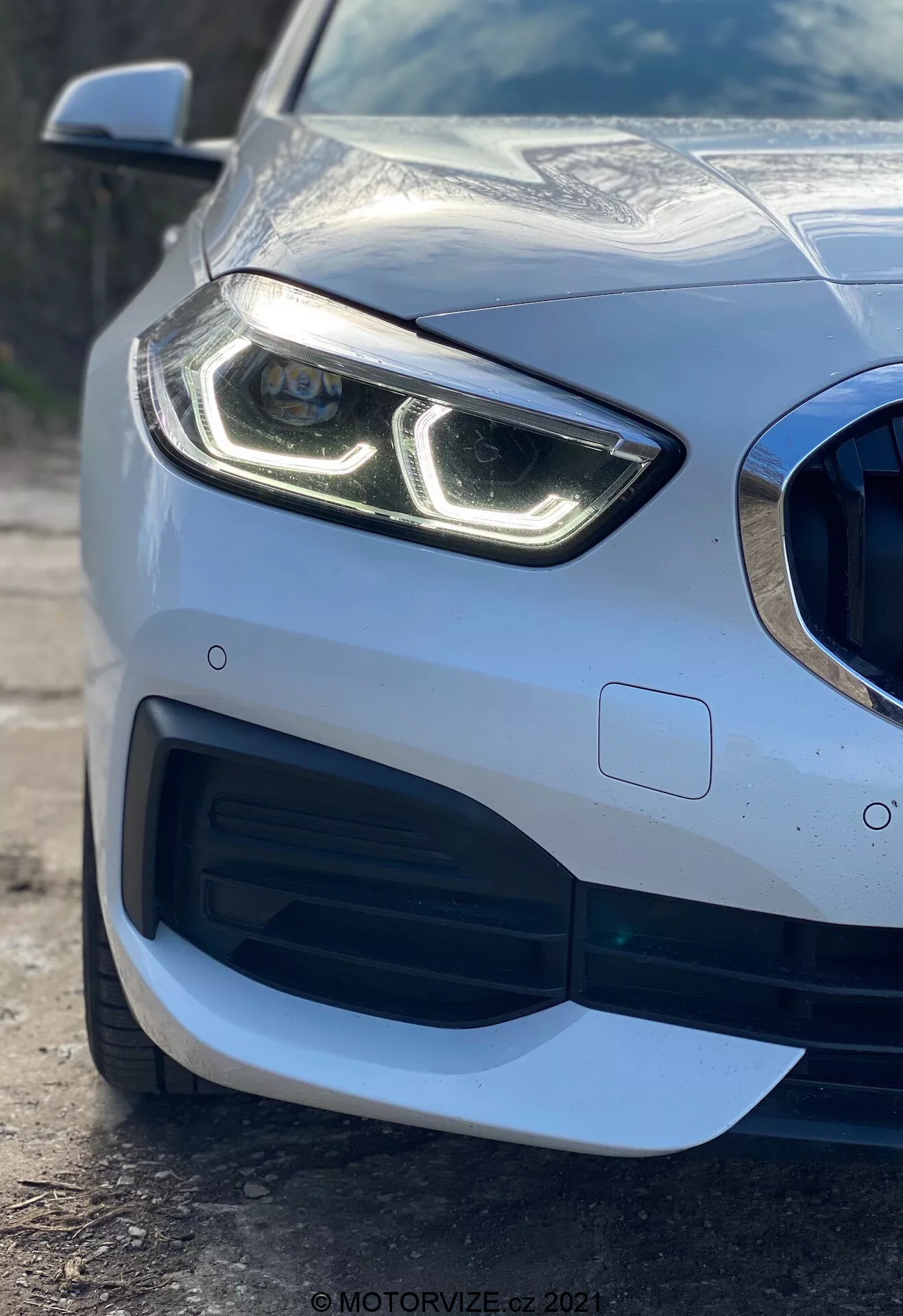 Közelkép egy 2019-es BMW 1-es sorozatú (BMW 116i) autó bal első részéről, kiemelve a modern, jellegzetes LED nappali menetfényes fényszórót, a méhsejt mintás hűtőrácsot nagy légbeömlővel, a kifinomult aerodinamikai kialakítást az első lökhárítón és a lökhárítóba integrált parkolószenzort, bemutatva a prémium metálfényezést és a visszaverő karosszériát.