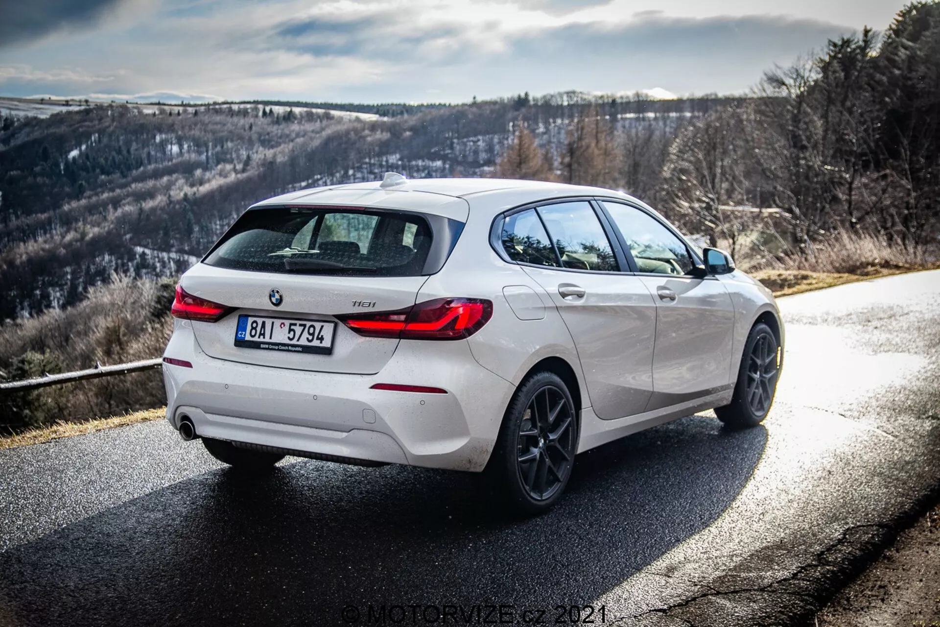 Dinamikus háromnegyedes hátsó nézet egy fehér BMW 116i 2019-ről, amely nappali fényben jobboldali és hátsó részét mutatja, tiszta napsugarakkal és árnyékokkal. A kép kiemeli a dizájn részleteit, beleértve a fekete kiemeléseket a hátsó diffúzoron, a keréktárcsákon és az oldalsó tükör burkolatokon, kiegészítve alufelnik és kettős kipufogórendszer, sportosabb változatot sugallva. Az út jobbra kanyarodik, hegyvidéki vagy dombos háttérrel, ideális autózási rajongók számára.
