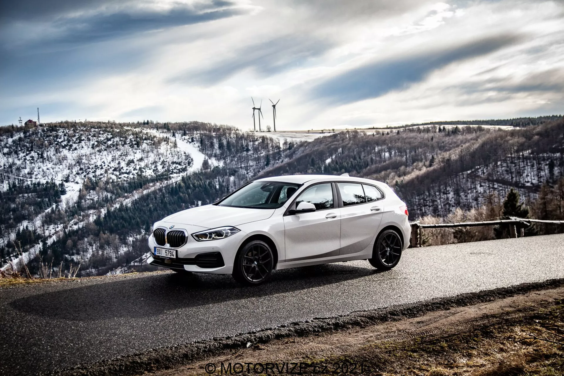 Egy fehér BMW ferdehátú autó, valószínűleg egy 2019-es BMW 1er-Reihe (BMW 116i), parkol egy ívelt aszfalt úton, melyet dombos, havas táj vesz körül szélkerekekkel. A fotó, mely elölről, oldalról készült, kiemeli az autó elülső rácsát, fényszóróit, aerodinamikai vonalait, és alumínium kerekeit a borús égbolt alatt.