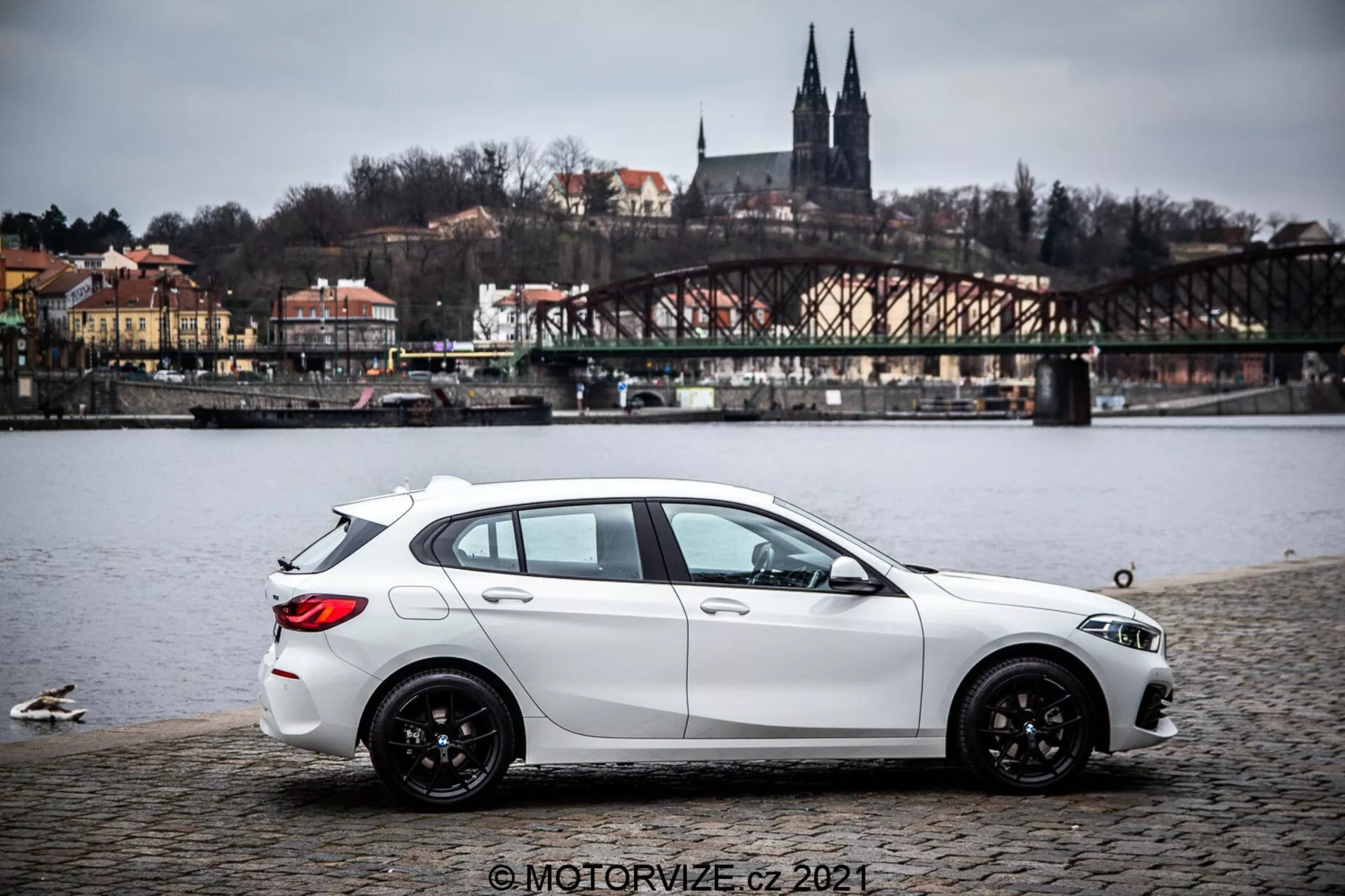Egy 2019-es BMW 1-es sorozatú (BMW-116i) fehér hatchback kocsi macskaköves felületen parkolva egy víztest mellett kacsákkal, oldalnézetből enyhén hátsó negyed perspektívával. Az autó dinamikus formázott karaktervonalakkal, lejtő tetővonallal, fekete többlüktű könnyűfém kerekekkel és látható fényszórókkal és oldalsó tükrökkel rendelkezik. A háttérben híd, dombon lévő épületek és építészeti elemek, amelyek európai környezetet sugallnak. A világítás szórt borús égbolt alatt, a kép pedig tartalmaz egy © MOTORVIZE.cz 2021 vízjelet.