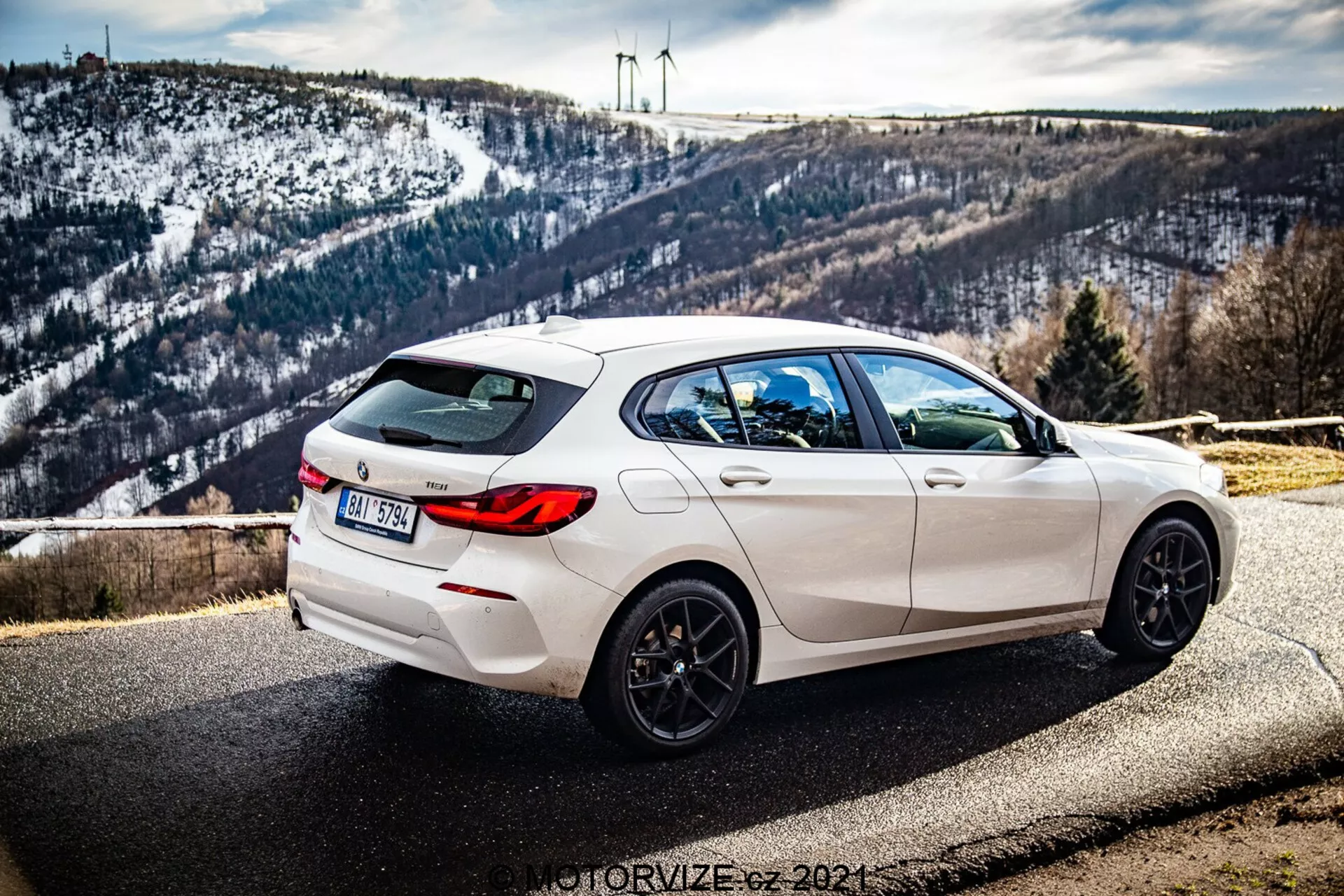 A képen egy fehér 2019-es BMW 1-es sorozat (BMW-116i) parkol az útszélen egy festői háttér előtt, amely egy dombos, hóval borított tájat és turbinákat tartalmaz a láthatáron. A fényképet háromnegyed hátsó szögből készítették, kiemelve a jármű jobb oldalát. A nézet bemutatja az elegáns formaterv vonalait, hátsó spoilerét, öt- küllős fekete könnyűfém kerekeit, kettős kipufogóit és a hátsó sötétített üveget. Az autón hátsó ablaktörlő és beépített cápauszony antenna található a tetőn. A mélységélesség azért tartja élesen az autót, mert a nappali fény kiemeli sportos esztétikáját, miközben a háttér finoman részletezett marad.