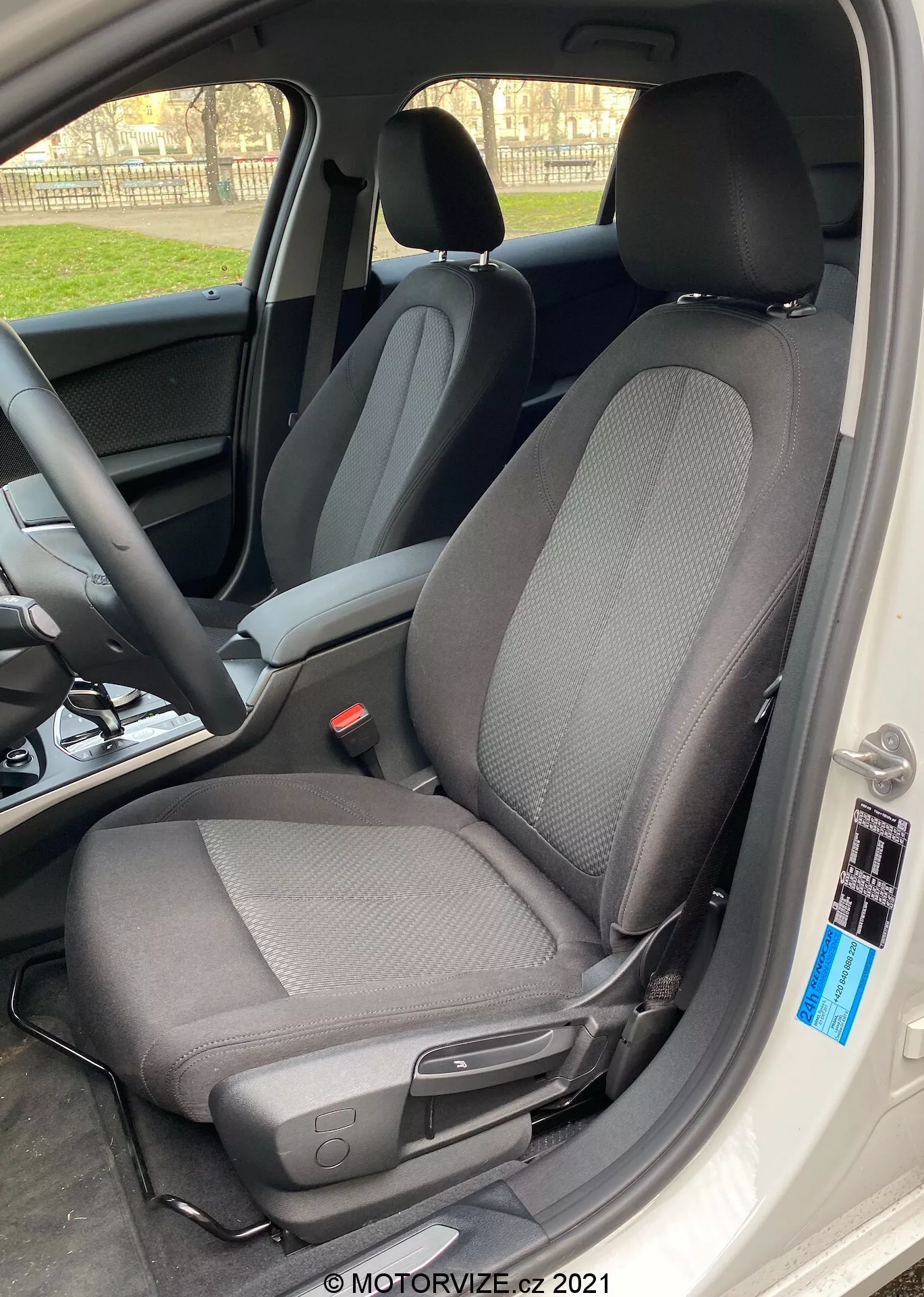 Közelkép a BMW 1er-Reihe 2019 (BMW-116i) belső teréről, amely elsősorban a vezetőülésre összpontosít modern, kétszínű szövettel, amely túlnyomórészt fekete, világosszürke mintákkal, oldalirányból, nyitott autóajtóval. Az üléshez fejhallgató tartozik, amely a fény felé dőlve sziluett hatást kelt a szélein, és tartalmaz egy kart a kézi előre vagy hátra állításhoz, valamint egy kis fekete gombot az autó belsejének szélén. A természetes nappali fény kiemeli az ülés éles fókuszát, míg a háttér elmosódik, megmutatva a kormánykereket és a műszerfalat.