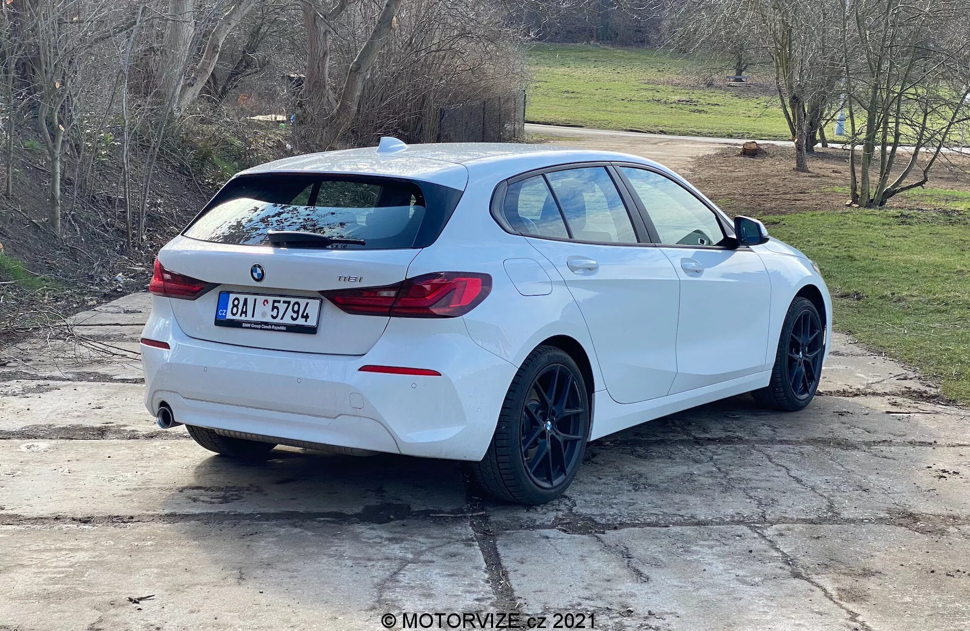 Háromnegyedes hátsó nézet egy fehér BMW 1-es sorozatú (2019-es BMW 116i) ferdehátú autóról, amely nappali fényben parkol a szabadban. Az autó oldalprofilja és hátulja jól látható, BMW emblémával, modelljelzéssel, jellegzetes csomagtérajtó körüli hátsó lámpákkal, hátsó spoilerral, fekete felnikkel, ferdehátú karosszéria kialakítással, dupla kipufogócsövekkel, hátsó ablaktörlővel, enyhén sötétített ablakokkal és integrált irányjelzővel ellátott visszapillantó tükrökkel. Az autó egy betonfelületen helyezkedik el, melyet fűfoltok és vidéki vagy külvárosi környezet övez.