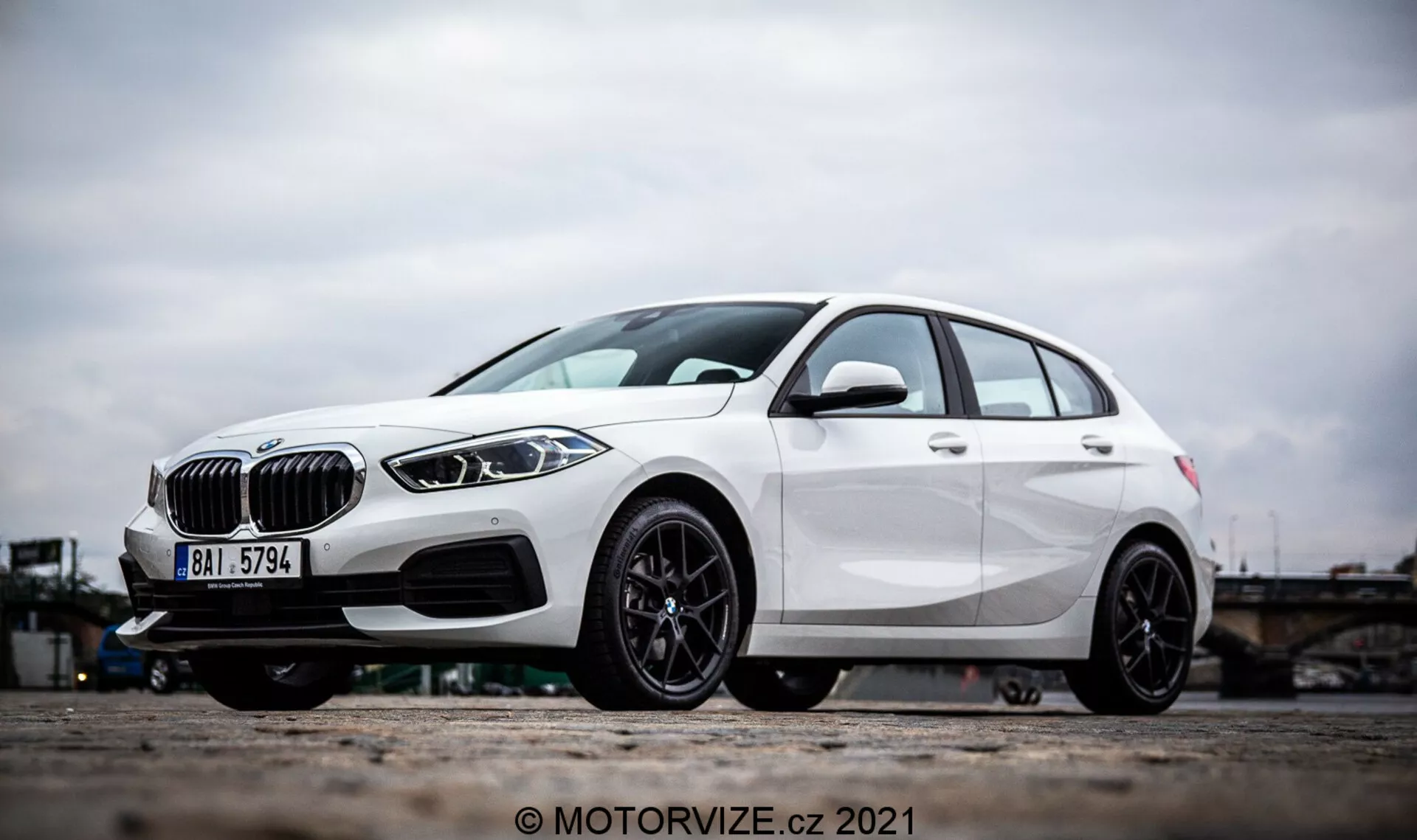 Egy fehér BMW 1-es sorozat 2019 (BMW 116i) ferde oldalnézete, amely kiemeli a jellegzetes tervezési elemeket, mint a vesekarima alakú hűtőrács, elegáns LED fényszórók és első lökhárító légbeömlőnyílásokkal és esetleges radarszenzorokkal a vezetői segédrendszerekhez. A fotó a könnyűfém kerekekre és a jármű alsó részére összpontosít, kiemelve a sportos, fekete, többlankás dizájnt és a teljesítményorientált tárcsaféket. A jellegadó vonalak és az aerodinamikus formázás a hatchbacken tovább hangsúlyozzák modern és dinamikus külsejét.