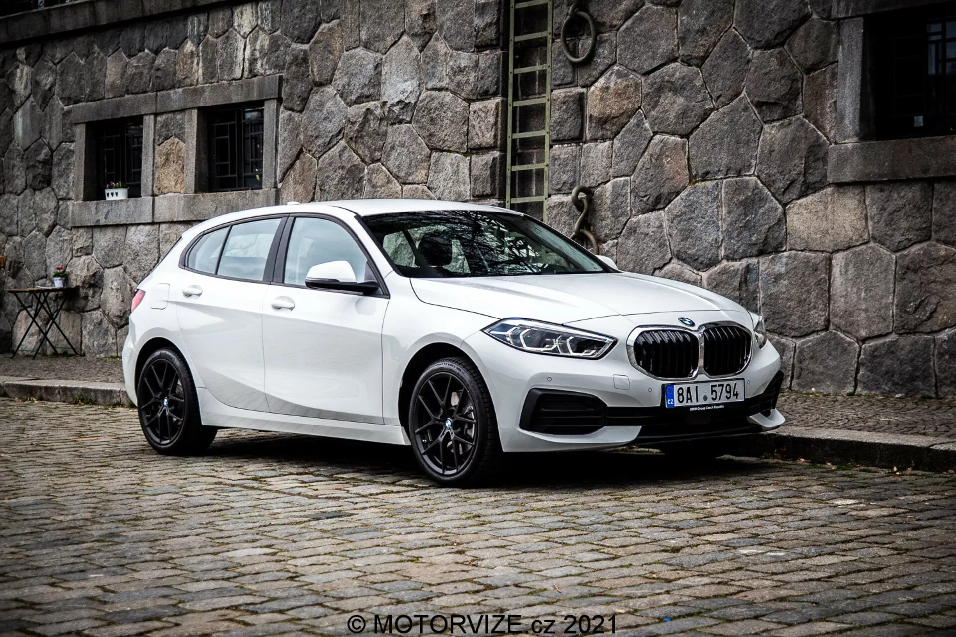 Egy fehér BMW 1er-Reihe 2019 (BMW 116i) hatchback háromnegyedes frontális szögből nézve, megmutatva a jármű bal oldalát. A fotó kissé alacsonyabb szögből készült, kiemelve az autó állását és tervezési vonalait. Egy macskaköves utcán helyezkedik el, háttérben egy régi kőépülettel, klasszikus városi jelenetet teremtve. Az első részen jól látható a vese alakú hűtőrács és a BMW embléma, elegáns szögletes LED fényszórókkal szegélyezve. Az autó többküllős fekete alufelniket, lejtős tetővonalat, karosszéria színű oldalsó tükröket integrált irányjelzőkkel és sötétített ablakokat kínál a hátsó utasok számára.