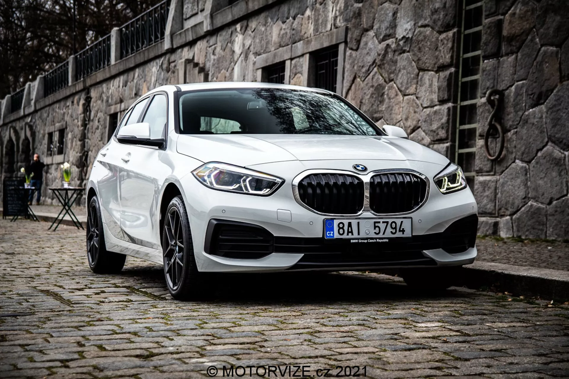 Modern fehér BMW 1-es széria (BMW 116i) 2019-es modell, jellegzetes hűtőrácsával, kettős kör alakú LED nappali menetfényeivel és sportos első lökhárító kialakításával, macskaköves felületen parkol egy történelmi európai környezetben, háttérben kőfallal és szabadtéri kávézóbútorokkal.