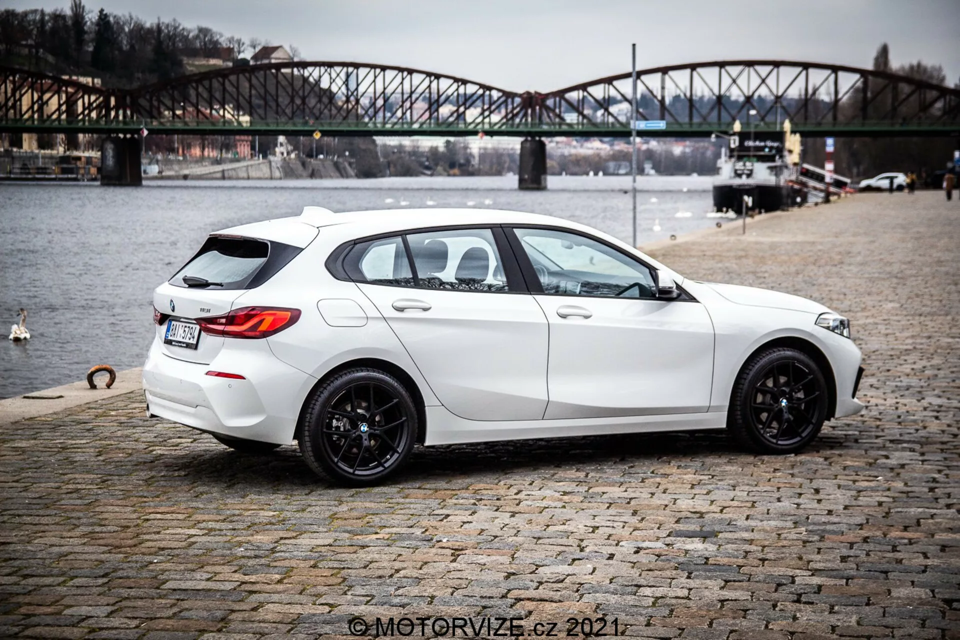 Kép egy fehér 2019-es BMW 1er-Reihe 116i ferdehátú autóról, hátsó háromnegyedes nézetből, kiemelve elegáns, modern formatervezését sportos megjelenéssel, fekete többrészű könnyűfém keréktárcsákkal, enyhén sötétített ablakokkal és praktikus funkciókkal, mint a hátsó ablak jégtelenítő és ablaktörlő. Az autó éles vonalakat és kontúrokat, kettős kipufogórendszert, finom hátsó tetőspoilert, tolatókamerát és parkolószenzorokat mutat be. Macskaköves felületen parkol egy víztest közelében, háttérben egy városi híddal, ami kültéri életmódkontextust sugall.