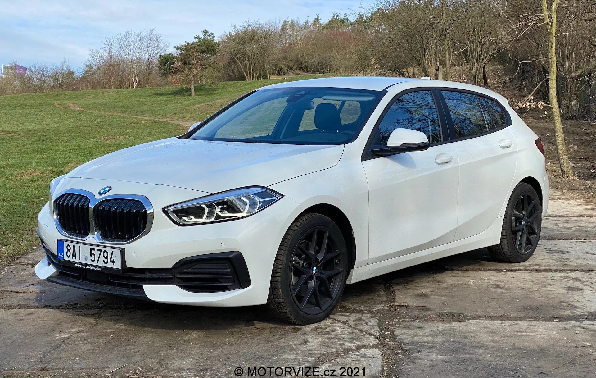A képen egy modern, fehér BMW 1-es sorozat (BMW 116i) ferdehátú látható, szabadtéri, füves háttérrel. A vezető oldalát előnyben részesítő, elölről háromnegyed nézetből rögzítve, bemutatja a jármű jellegzetes veserácsát, elegáns, szögletes LED-fényszóróit, fekete könnyűfém kerekeit, jól definiált oldalíveit és az oldalsó tükrökbe integrált irányjelzőket. Az első lökhárító parkolásérzékelőkkel van felszerelve, míg a panorámás üvegtető és az esetleges elülső kamera feltételezhető. Az autó kőburkolatú területen parkol, és a kép nagy részét elfoglalja, ami modern járműtervezést és fejlett technológiai jellemzőket sugall.