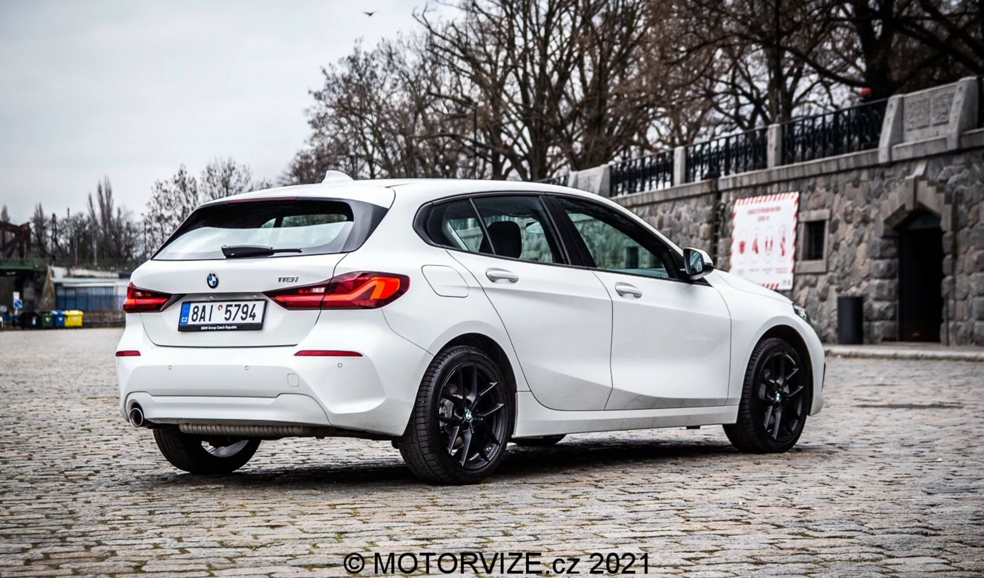 Egy magas minőségű fénykép egy 2019-es fehér BMW 1 Series 128ti hatchback-ről, háromnegyedes, hátsó szögből fényképezve, kiemelve a jobb oldalát. Az autó egy macskaköves járdán parkol, háttérben kövekkel kirakott fal, kiemelve sportos kialakítását hátsó tetőszpoilerrel, kettős kipufogórendszerrel, hátsó diffúzorral, LED-es hátsó lámpákkal, sötétített ablakokkal és sötét színű könnyűfém felnikkel. A szórt megvilágítás, az éles árnyékok hiánya és a tiszta részletek felhős környezetet sugallnak, autentikus ábrázolást nyújtva a jármű színéről és jellemzőiről. A képen látható a 'MotorVize.cz 2021' vízjel.
