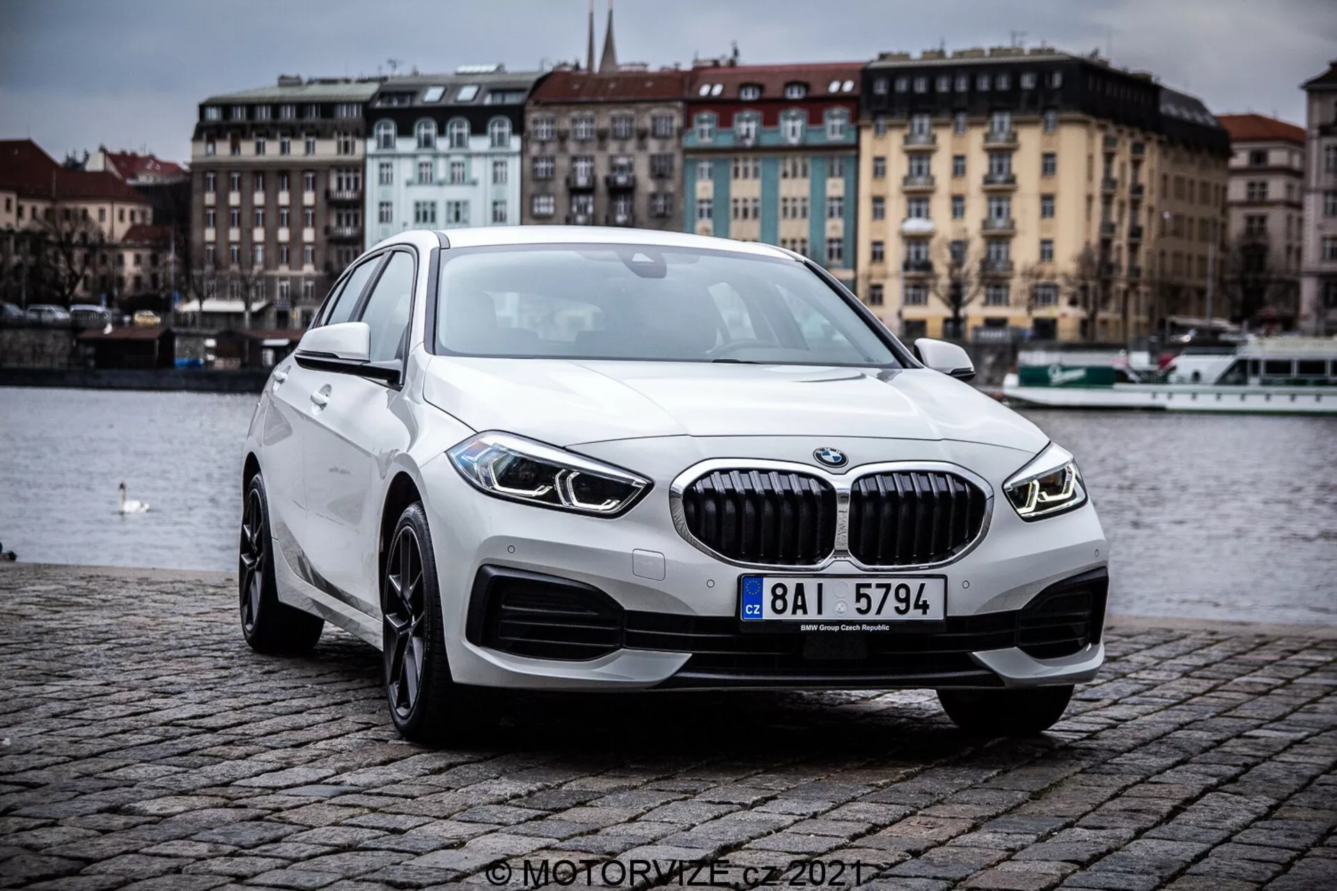 Egy 2019-es BMW 1er-Reihe (BMW 116i) képe, elöl háromnegyed perspektívából, kiemelve a modern luxus sedan kontúrjait és formatervezési elemeit. Az autó kint van egy macskaköves töltésen, egy víztest mellett, felhős ég alatt. Jellegzetes elemek közé tartozik a függőleges lamellákkal ellátott hűtőrács, elegáns LED fényszórók, integrált ködlámpák, könnyűfém kerekek és aerodinamikus profil.