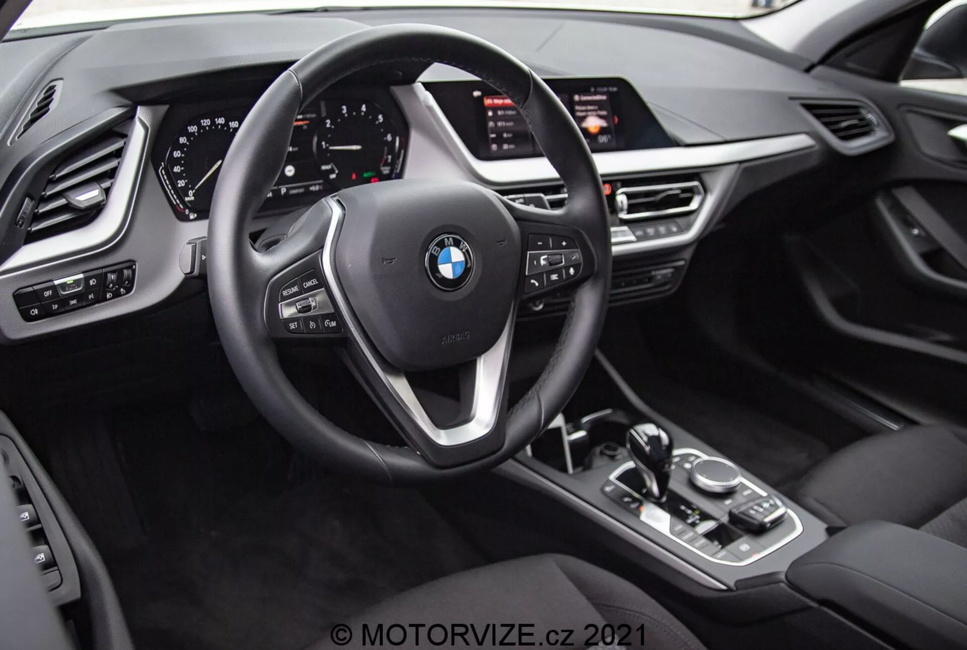 A BMW 1er-reihe 2019 (BMW 116i) belső nézete a vezető perspektívájából. A képen látható a BMW embléma a háromküllős kormánykeréken, amely többfunkciós gombokkal és váltófülekkel van felszerelve. A kormánykerék mögött egy teljesen digitális műszercsoport látható, világító számlapokkal és különböző információs kijelzőkkel. A középkonzolon egy automata sebességváltó kar, iDrive vezérlő gomb, vezetési módok gombjai és egy nagy, a vezető felé döntött infotainment képernyő található. A belső tér bőrhatású textúrákkal, fémes elemekkel és további vezérlőkkel rendelkezik a fényszórók, az ablakok és a visszapillantó tükrök beállításához, valamint memóriafunkcióval a vezetőülés számára.