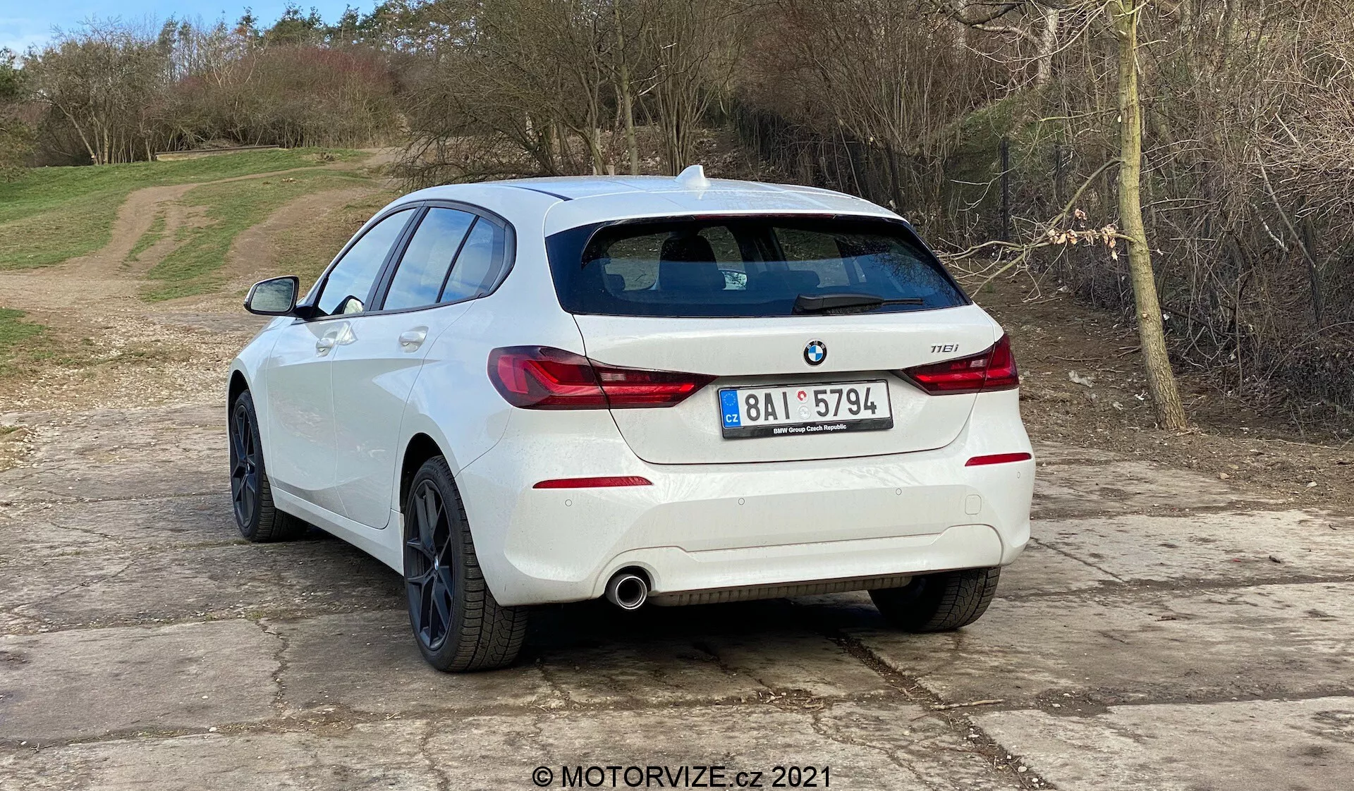 A képen egy fehér BMW 1er-reihe 2019 (BMW 116i) hatchback látható háromnegyed hátulnézeti szögből. A jármű hátsó része, beleértve a vízszintesen orientált hátsó lámpákat, a kettős kipufogórendszert és a hátsó lökhárítóba integrált parkolószenzorokat, jól látható. A BMW embléma és a modell jelzése a hátsó ajtón található. Az autó egy kavicsos felületen van elhelyezve, természetes, vidéki háttérrel. A kép autós témákat sugall, például járműtervezést, kortárs autómodelleket, BMW-specifikus tartalmakat, kipufogórendszereket és parkolási asszisztens technológiát. A MOTORVIZE.cz 2021 vízjel jelenléte autós áttekintés vagy publikáció eredetére utal.