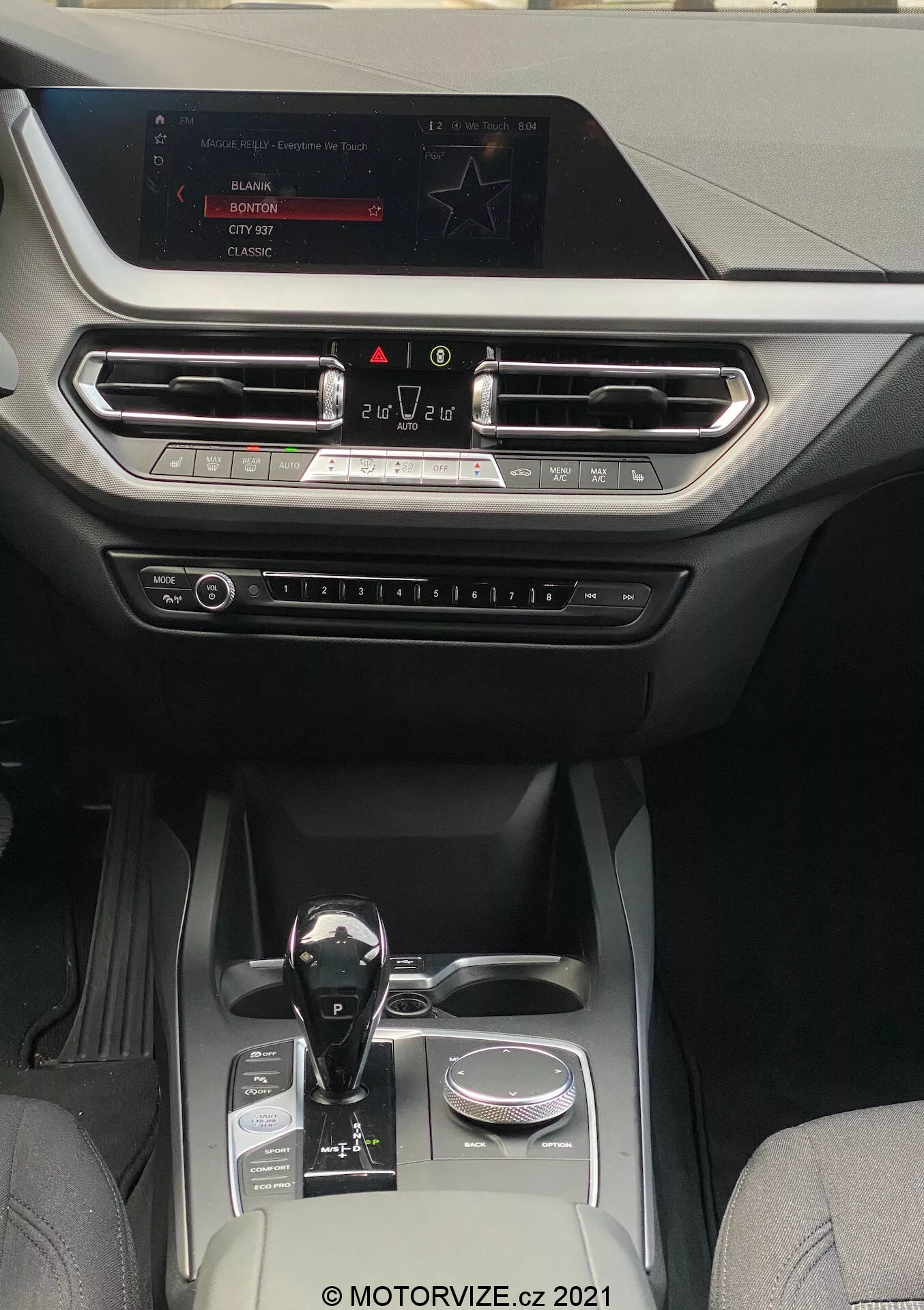 A 2019-es BMW 1er-reihe (BMW-116i) belső nézete, amely egy modern és luxus kabint mutat be, a középkonzolra és a műszerfalra összpontosítva. A perspektíva a hátsó ülésekről néz előre, az infotainment képernyő felé, amely a műszerfal tetején van elhelyezve. A kijelző rádió, média és autóbeállítási lehetőségeket mutat. A képernyő alatt vízszintesen igazított szellőzők találhatók fémes díszítéssel és egy dekoratív léccel, amely a műszerfal teljes szélességében végigfut. A szellőzők alatt fizikai gombok és tekerők láthatók a klímaberendezés és más beállítások szabályozására. Az automata váltókar fényes fekete felülettel és fémes díszítéssel rendelkezik, egy forgókapcsoló mellett helyezkedik el. A középkonzol két pohártartót, egy kis tárolórekeszt és egy ezüst díszítőelemet tartalmaz, ami jól felszerelt kabint jelez, amely bőrt és magas minőségű szintetikus szöveteket kombinál, és közepes és magas kategóriájú járművekhez alkalmas, amelyek a technológiára és a kényelemre összpontosítanak.