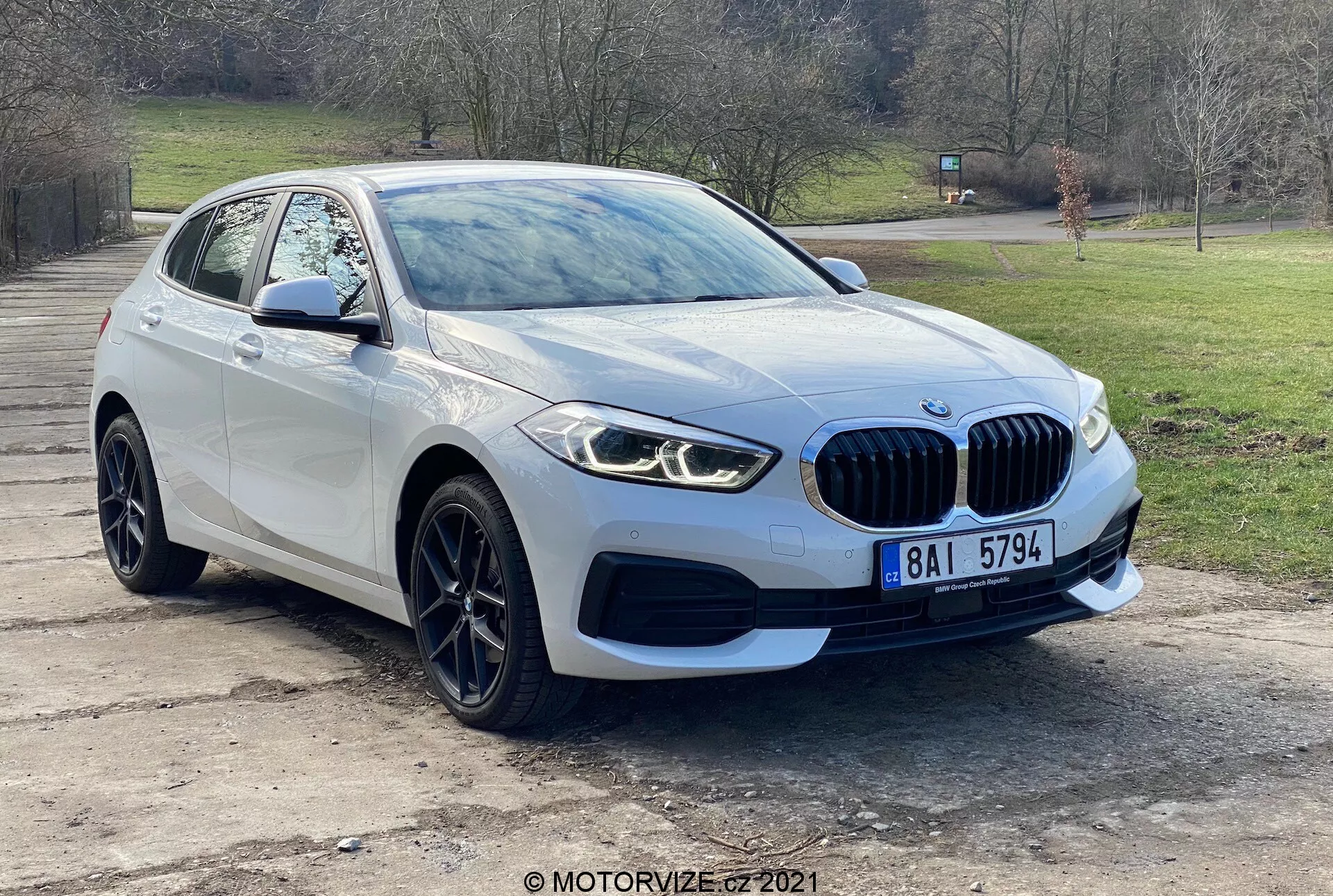 Modern BMW 116i jármű a 2019-es 1-es sorozatból, háromnegyedes elülső perspektívából fotózva, bemutatva az emblémával ellátott első rácsot, elegáns LED fényszórókat, ködlámpát, első lökhárítót és a szélvédőig vezető motorháztető aerodinamikus vonalait. A vezető oldali kerék és sárvédő, valamint a beépített irányjelzőkkel ellátott oldalsó tükör kiemelik a jármű fejlett dizájnját és parkolásérzékelő technológiáját. Külső felvétel a fák és a fű hátterével, az autó alufelnikkel és jellegzetes veserácsokkal rendelkezik, jelezve a luxust és a teljesítményt.