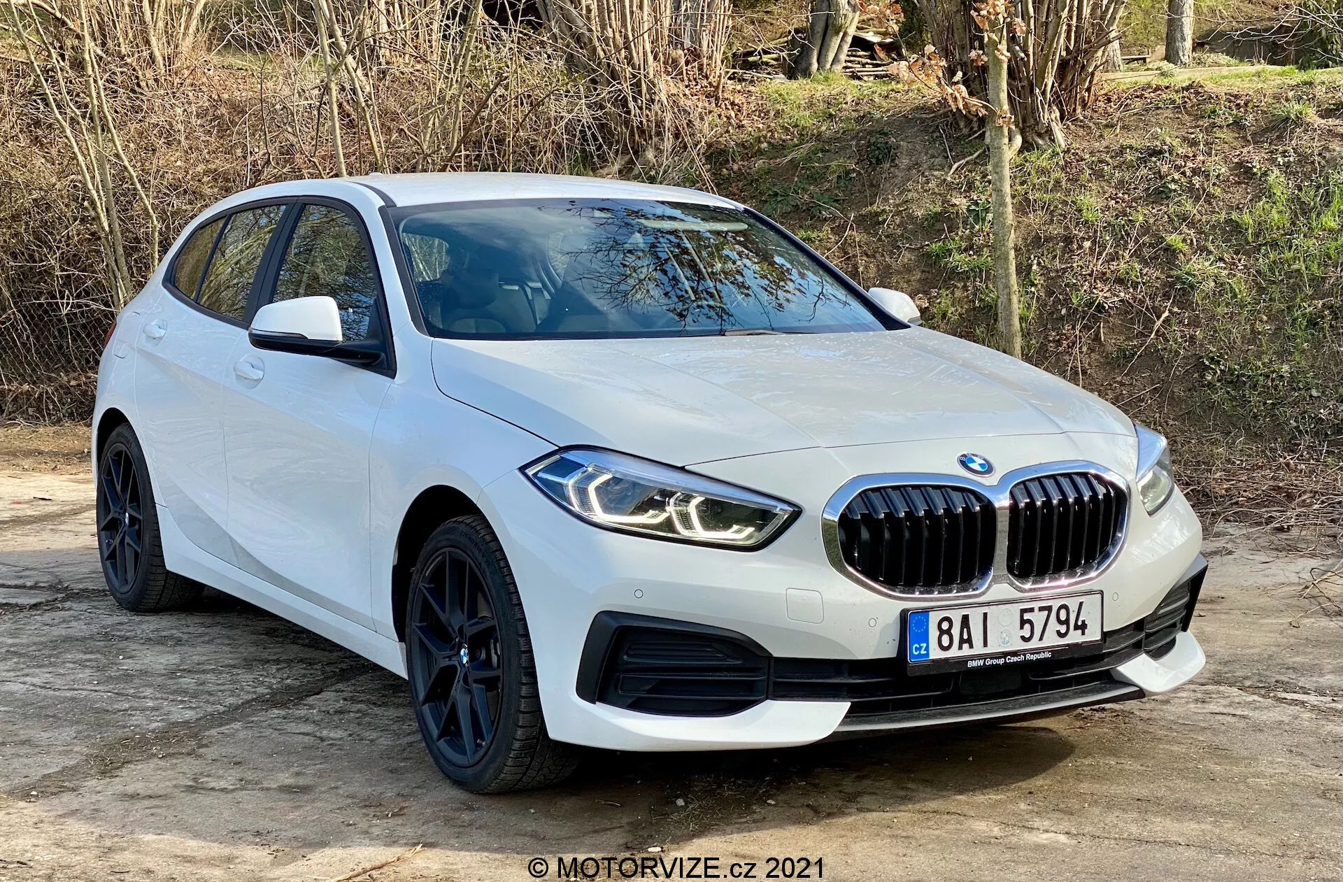 2019-es BMW 116i 1-es sorozat, fehér színben, frontális háromnegyedes szögből fényképezve, amely bemutatja a prominens vese alakú hűtőrácsot, az éles szögű LED fényszórókat, az elegáns fekete kerekeket alacsony profilú gumiabroncsokkal és az áramvonalas karosszéria vonalait. Sík felületen helyezkedik el, természetes növényzet háttérrel, amely kiemeli az autó modern dizájnját és sportos jellemzőit, beleértve a parkolószenzorokat és az integrált irányjelzőket az oldalsó tükrökön.