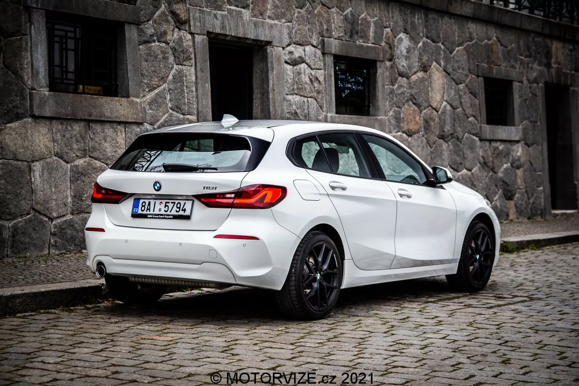 Közeli, talajszintű kép egy fehér 2019-es BMW 1er-sorozat 116i hatchbackról háromnegyed hátsó nézetből, mely jelentősen megjeleníti a LED-es kialakítású hátsó lámpákat, a hátsó szélvédőt és a sportos felniket. A látható jellemzők közé tartozik a hátsó lökhárító érzékelőkkel, amelyek valószínűleg parkolási segítséget jelentenek, egy kis tetőantenna, amely valószínűleg GPS vagy műholdas rádió számára, modelljelzés a csomagtartón, kettős kipufogórendszer, amely teljesítményt sugall, és sötétített ablakok a magánéletért vagy a kényelemért.
