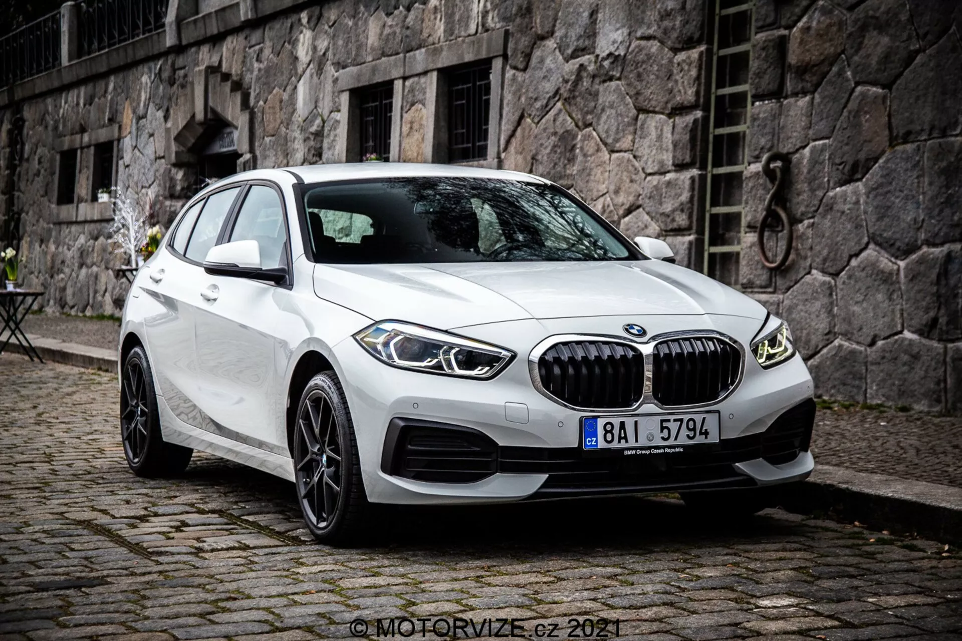 A kép egy 2019-es BMW 1-es szériát, konkrétan a BMW 116i-t ábrázolja fehér színben. Az autó egy elülső háromnegyedes szögből van fényképezve, így jól látható az autó elülső része, az utasoldala és a kerekek részletes kialakítása. A fotó kissé megemelt nézőpontból készült, ami arra utal, hogy a fotós állt vagy tripoddal dolgozott. Az autó álló helyzetben van, az első kerekek jobbra fordítva, ami tiszta betekintést nyújt a felnik és a gumiabroncs oldalsó profiljának kialakításába. A jellemzők közé tartozik a kiemelkedő elülső rács függőleges lécekkel, az áramvonalas vonalak a motorháztetőn és az oldalsó paneleken, valamint a vékony LED-fényszórók és a lökhárítón lévő légbeömlők, amelyek parkolásérzékelőket is tartalmazhatnak. Az oldal tükrök beépített irányjelzőkkel és a keret nélküli ablakok a prémium esztétikát hangsúlyozzák. A többrészes könnyűfém felnik egy magasabb felszereltségi szintet vagy egy opcionális fejlesztést sugallnak. A meredek hátsó ablak kiemeli a ferdehátú profilját. A jármű macskaköves úton parkolt egy városi vagy történelmi környezetben, egy kőfalas épülettel és egy kis bisztró stílussal rendelkező asztallal a háttérben. A kép éles és részletes, természetes felhős napfényben készült.