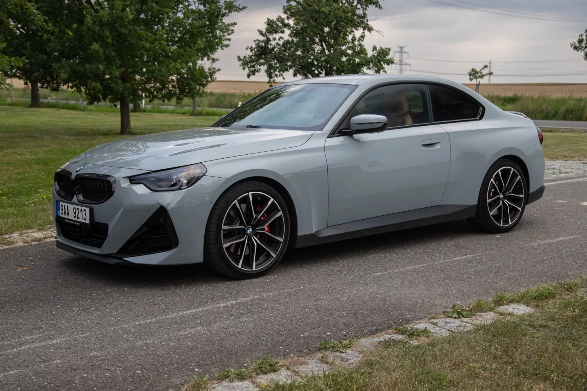A kép egy 2022-es BMW 2 Series Coupe-t ábrázol egy háromnegyedes elülső szögből, kiemelve az elegáns és sportos külső dizájnt, nagy elülső rácsozattal és szögletes LED fényszórókkal. A sportos kupé, matt világosszürkére festve, többrugós könnyűfém kerekekkel, nagy teljesítményű fékrendszerrel piros féknyergekkel és szoborszerű testvonalakkal rendelkezik az aerodinamikus profil érdekében. Az autó egy kövezett úton parkol, természetes tájjal és mezővel a háttérben, nappali fényviszonyok között.