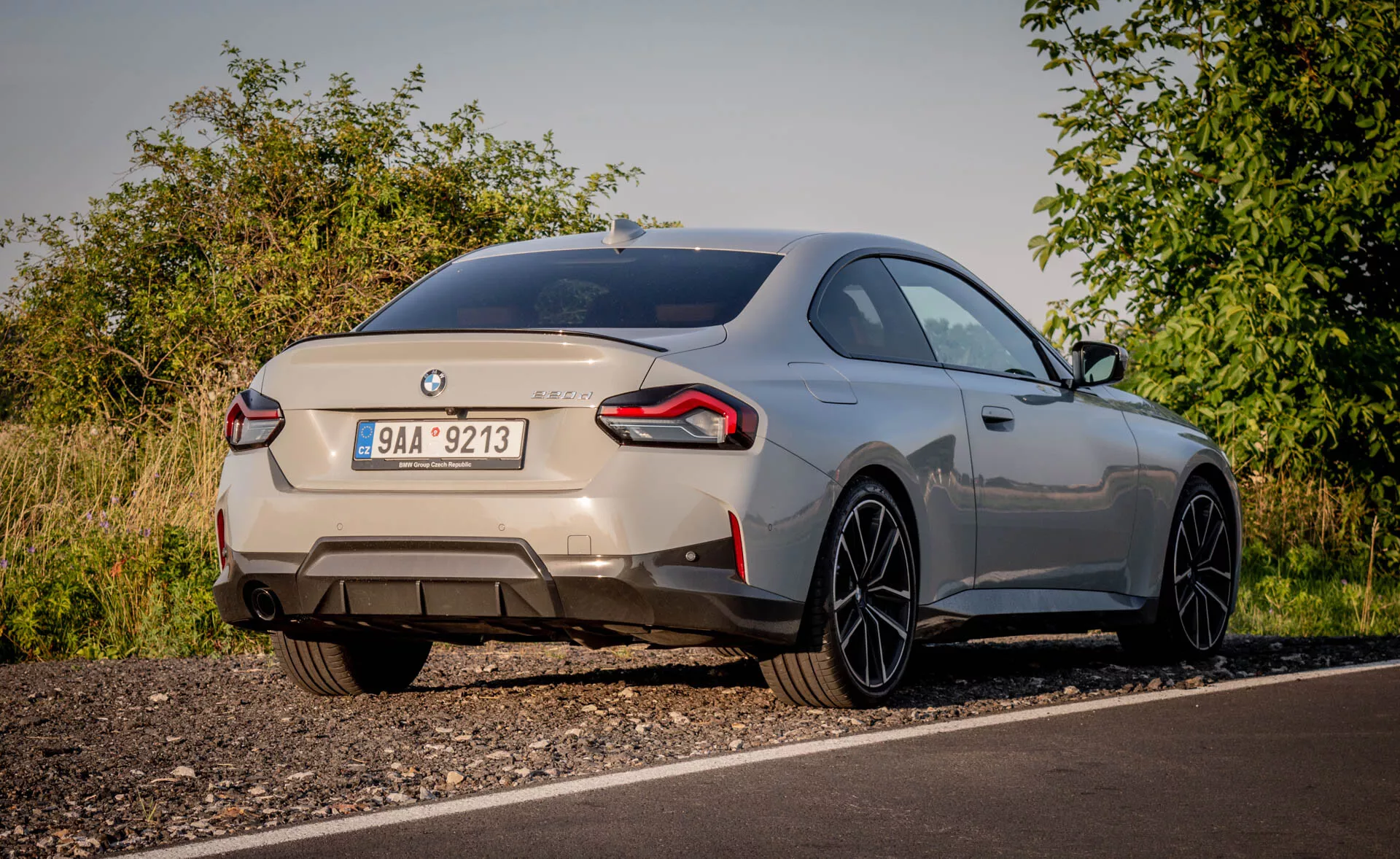 BMW 2er-reihe 2022 (2-es sorozat kupé) sportautó hátsó nézete, alacsony szögtől nézve. Az autó a természetes lombdal körülvett, vidéki környezetben egy út szélén parkol, lágy fényviszonyok között. Az egyedi emblémák azonosítják a márkát és a modellt, bemutatva a hátsó részt, beleértve a csomagtartó fedelet, a hátsó lökhárítót és az oldalra kiterjedő hátsó lámpákat. A kettős kipufogócsövek erős motort jeleznek. Az autó hátsó részén egy kicsi, integrált hátsó spoiler és nagy, vékony küllős könnyűfém felnik találhatók, amelyek aerodinamikai stílust és teljesítményt sugallnak. A fémes festés és a simán lejtő tetővonal prémium megjelenést kölcsönöz a járműnek. A látható modern funkciók közé tartozhatnak a LED-es hátsó lámpák, a parkolásérzékelők és a tolatókamera.