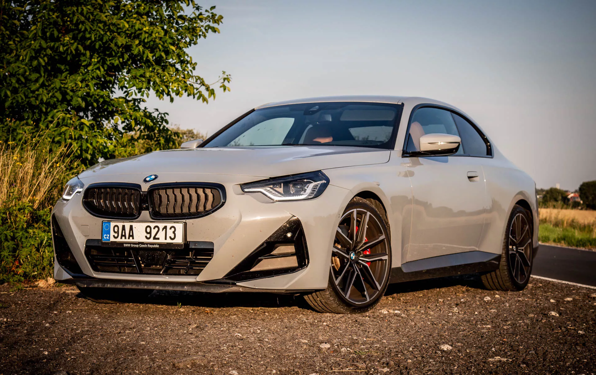 A kép a 2022-es BMW 2-es sorozat modern sportkupéját ábrázolja. Az autó az út szélén, nappal parkol, ferde szögből nézve, bal oldalról, kiemelve a vezető oldalát. A jellemzői közé tartozik a karcsú, aerodinamikus dizájn, a nagy, többrudas könnyűfém felnik sötét felülettel, kontrasztos féknyergek, egy jellegzetes hűtőrács, amit keskeny, agresszív LED fényszórók szegélyeznek, egy elülső kamera a rendszámtábla fölött, nagy légbeömlők az első lökhárítóban, világos metálfestés, integrált irányjelzőkkel ellátott oldalsó tükrök és egy részleges kilátás a panorámatetőre. Az autó közel van a kamerához, háttérben vidéki vagy külvárosi jelenettel, meleg világítással és árnyékokkal, amelyek alacsonyan lévő napot jeleznek reggel vagy késő délután.