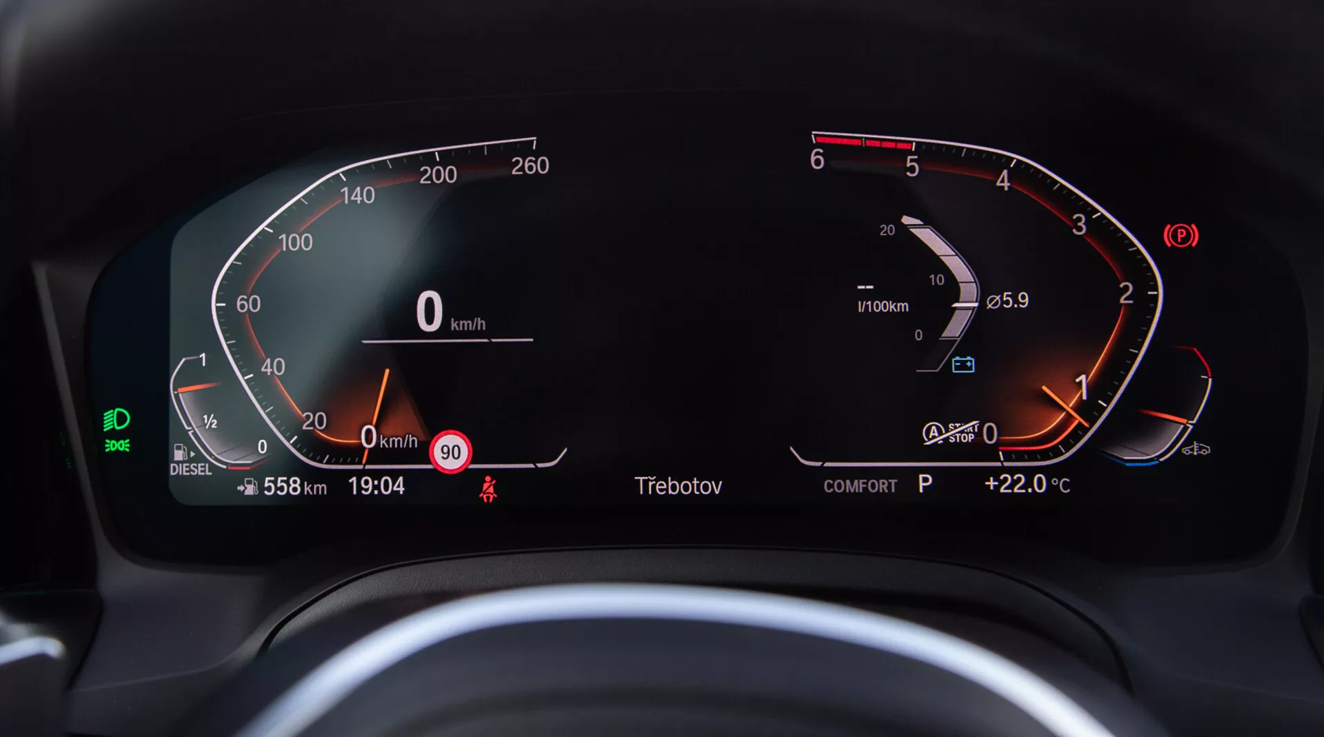 Közeli kép egy BMW 2 Series Coupe 2022 digitális műszerfaláról, amely a vezetőülésből látható. A bal oldali félköríves digitális sebességmérő 0 és 260 km/h között mozog, jelezve, hogy a jármű álló helyzetben van. A jobb oldali fordulatszámmérő éppen 0 RPM fölött mutatja a motort alapjáraton. A fordulatszámmérőbe integrált digitális üzemanyagszint-jelző majdnem tele van, és a motor hőmérséklete normál tartományban van. A központi digitális kijelzőn az össz-kilométer-számláló 558 km-t, sebesség 0 km/h, sebességhatár figyelmeztetés 90 km/h-nál, aktuális idő 19:04, 'P' sebességfokozat jelző Parkolásra és a külső hőmérséklet +22,0°C látható. Látható még egy zöld sávkövető asszisztens ikon, a 'COMFORT' mód kijelzése, diesel üzemanyag jelző és biztonsági öv emlékeztető is.