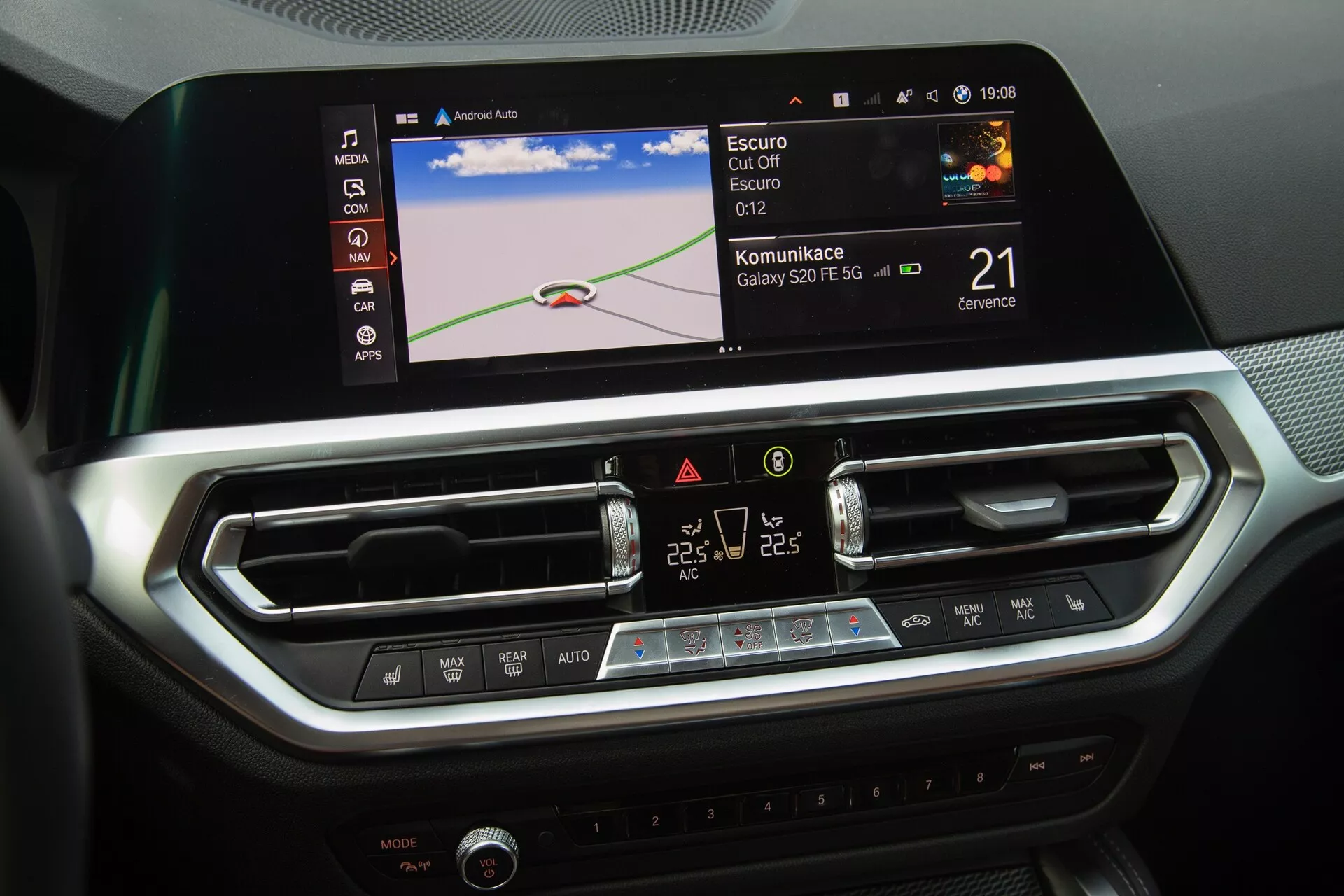 A BMW 2-Series Coupe 2022 középkonzolja van kiemelve, amely bemutatja a modern autó infotainment rendszerét és klímavezérlőit. A kép egy nagy színes érintőképernyőt ábrázol, amely navigációs térképet, Bluetooth vagy okostelefon integrációs ikonokat és órát jelenít meg. A képernyő alatt két központi légcsatorna állítható zsalukkal és egy elegáns panel érintőgombokkal és LCD kijelzővel a kétzónás klímavezérléshez. A belső tér, csiszolt fémes díszítésekkel és szénszálhoz hasonló textúrákkal, luxusos és technológia-orientált környezetet tükröz.