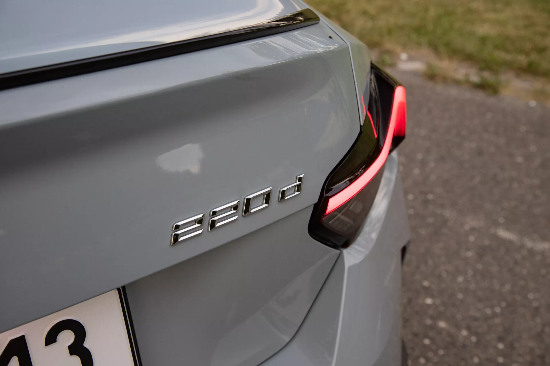 Egy BMW 530d hátsó jobb oldalának közeli részlete, kiemelve a '530d' modellmegjelölést és a modern, részben megvilágított LED hátsó lámpát. Az autó színe metalikus ezüst vagy szürke lágy fényvisszaverődéssel természetes fényben, ami estére vagy árnyékos környezetre utal. A logó króm vagy fém, kontrasztál a kocsi festésével és a csomagtérfedélen van elhelyezve. Alacsony mélységélességgel rendelkezik, ami kiemeli az '530d' logót és a közeli autótestet, miközben elhomályosítja a hátteret, alkalmas azoknak, akik érdeklődnek az autóipari technológia vagy járműtervezés iránt.