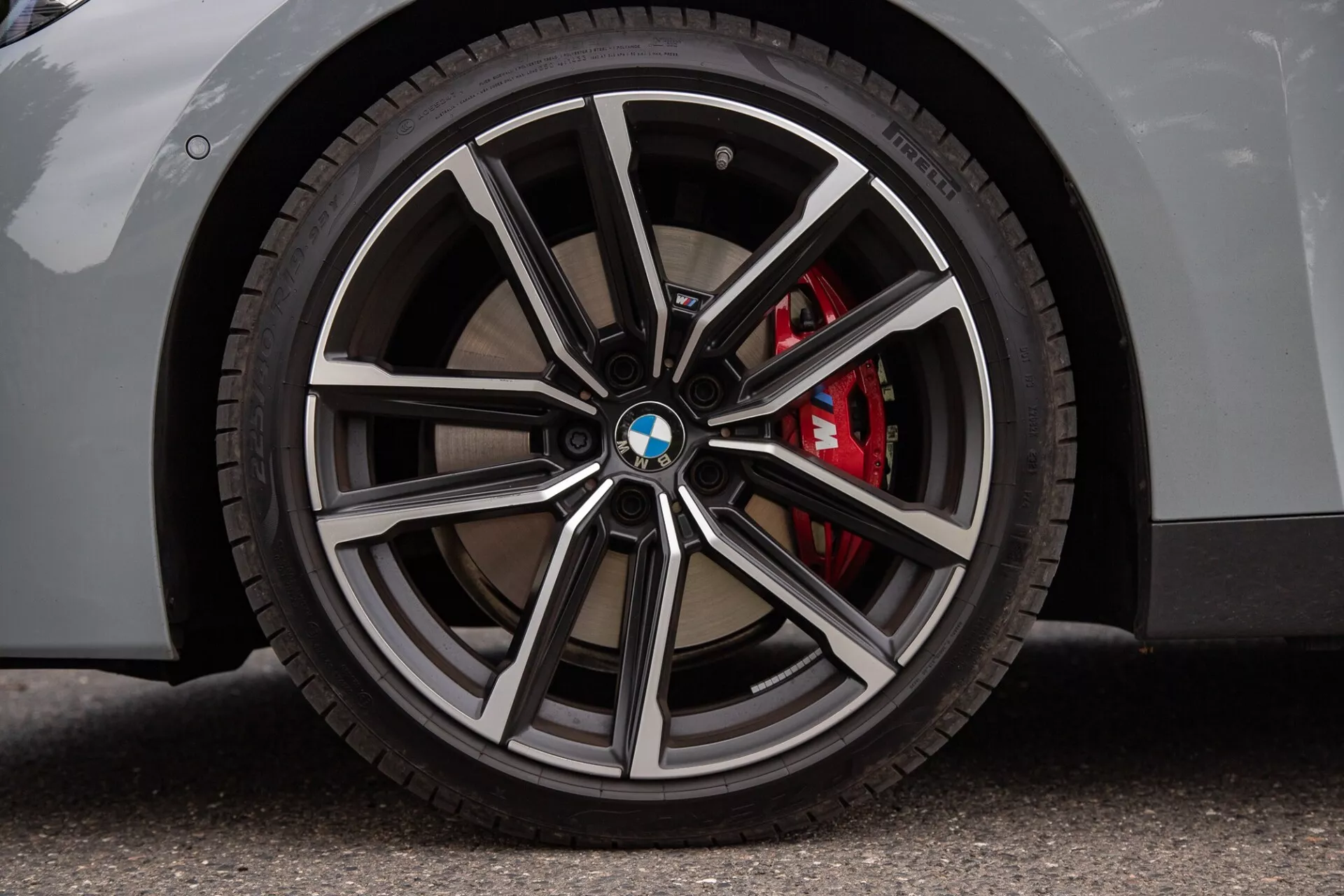 Közeli felvétel egy BMW 2-es Sorozat Coupé kerekéről, amely többküllős felnit mutat fényes és sötétebb fémes felületekkel, BMW logót a dísztárcsán, alacsony profilú gumit és piros teljesítmény féknyerget. Az aszfalton álló világos színű karosszéria hangsúlyozza a luxus és sportos jellegzetességeket.