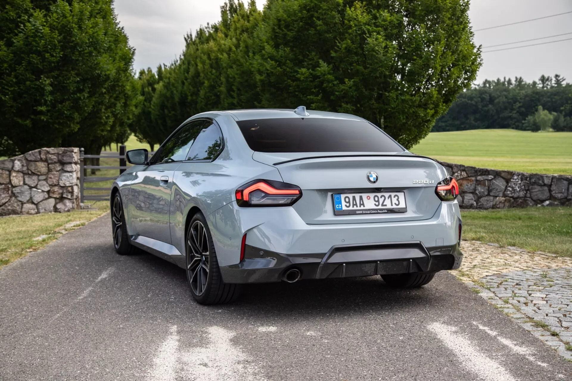 Kép a BMW 2er-Reihe 2022 2-Series Coupe-ról, amely hátsó háromnegyedes szögből lett megörökítve. Az autó, szinte átlós helyzetben, jellegzetes szögletes LED hátsó lámpákkal, két kipufogó csővel, többlengőkaros alufelnikkel és fényes metál fényezéssel rendelkezik. A hátsó kialakítás középen elhelyezett gyártói emblémát és a csomagtérajtó jobb alsó részén modell/motorkódot jelölő jelvényt mutat. A külső környezet burkolt útból vagy úttestből, füves területből, fákkal és részben látható kőfallal áll.