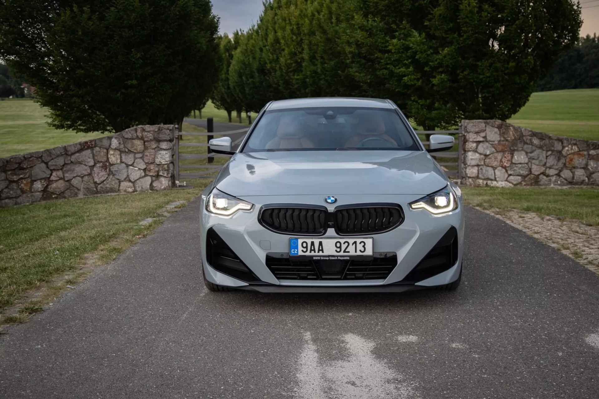 A 2022-es BMW 2 széria Coupé elölnézete egy parkosított, szabadtéri területen, amelyen kiemelik az autó jellegzetes márkarácsát, elegáns LED fényszóróit és aerodinamikailag formázott motorháztetejét, kiemelve modern dizájnját és technológiai jellemzőit, beleértve a fejlett vezetőtámogató rendszerekhez szükséges potenciális érzékelőket.