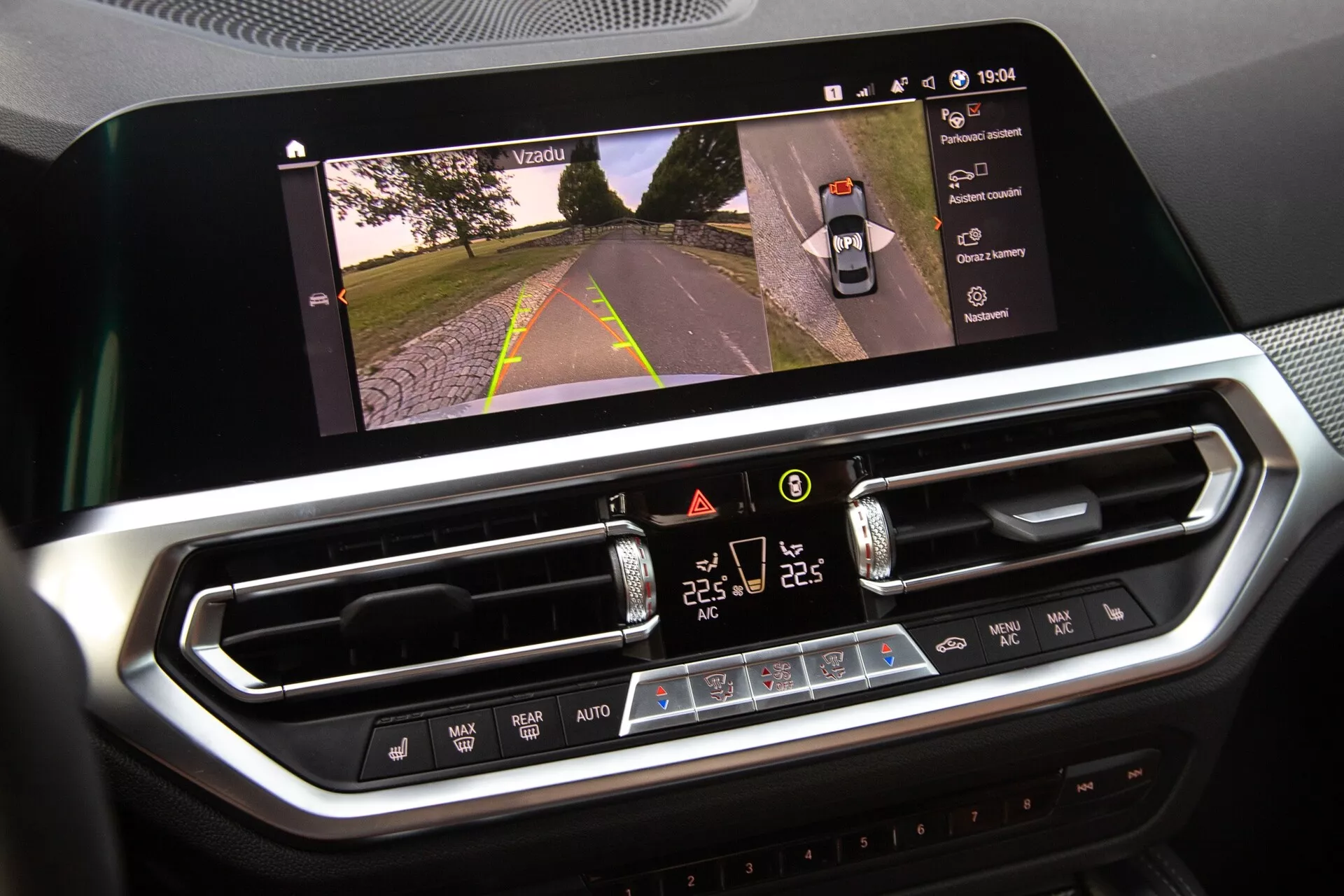 BMW 2er-Reihe 2022 belsőjének közeli felvétele, amely az infotainment kijelzőt és a klímavezérlő rendszert mutatja. A nagy, vízszintes érintőképernyő egy útnézetet jelenít meg, ami a kamera aktiválását jelzi, míg az alatta lévő digitális klímavezérlő felület kettőzónás hőmérséklet beállításokkal, ventilátor sebességgel és fizikai gombokkal rendelkezik. Modern, krómozott kialakítás prémium anyagokat sugall.