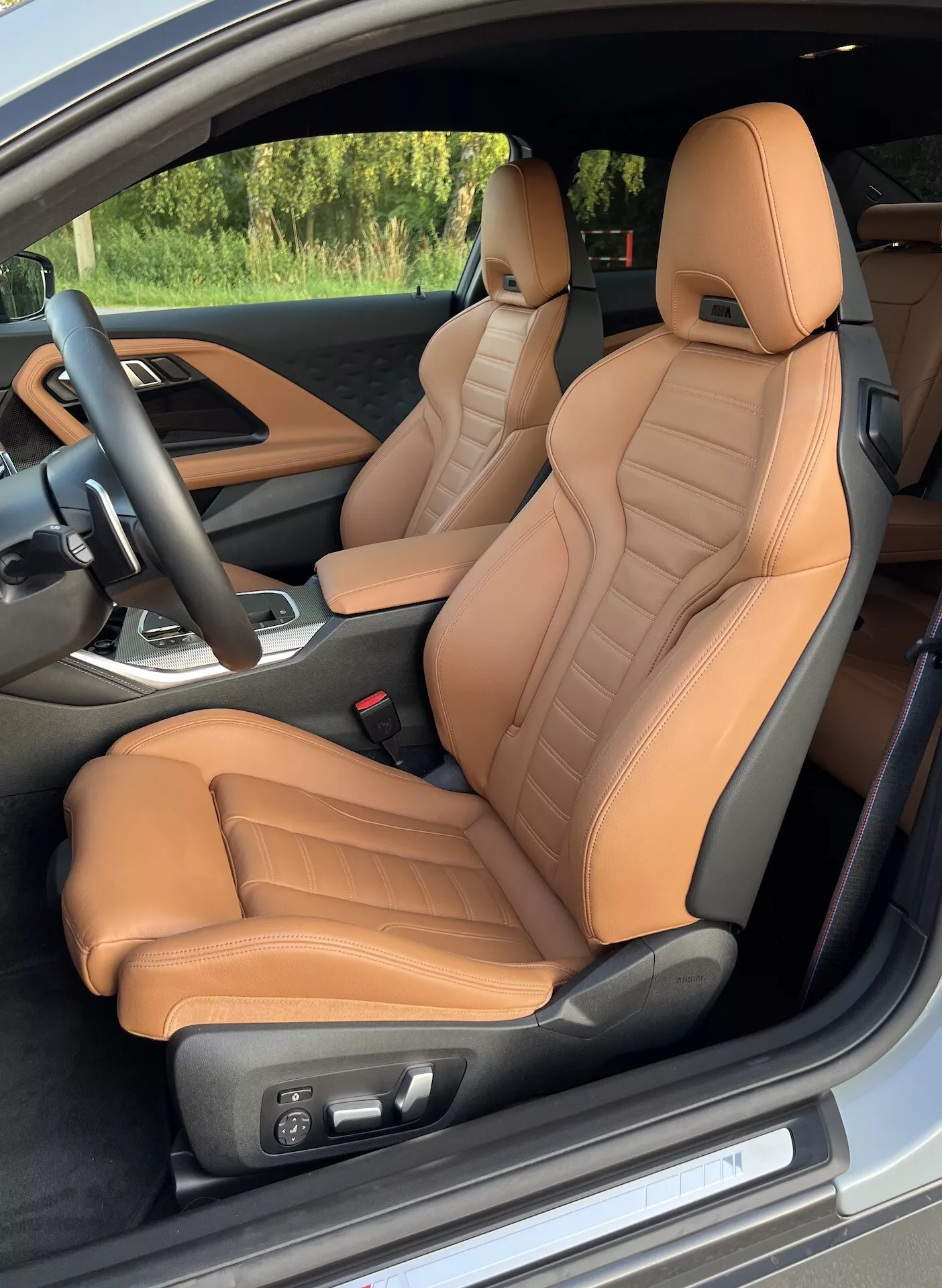 A 2022-es BMW 2-es sorozat kupé belső nézete a vezetőoldali ajtó felől, kiemelve a tan színű bőrrel kárpitozott első üléseket, steppelt részletekkel és ergonómiai támasztékkal. Az elektromos ülések állítása, a fekete kormánykerék integrált vezérlőkkel, valamint a sebességváltó kart is magában foglaló középkonzol részleges látványa luxust és teljesítményorientált funkciókat sugall.