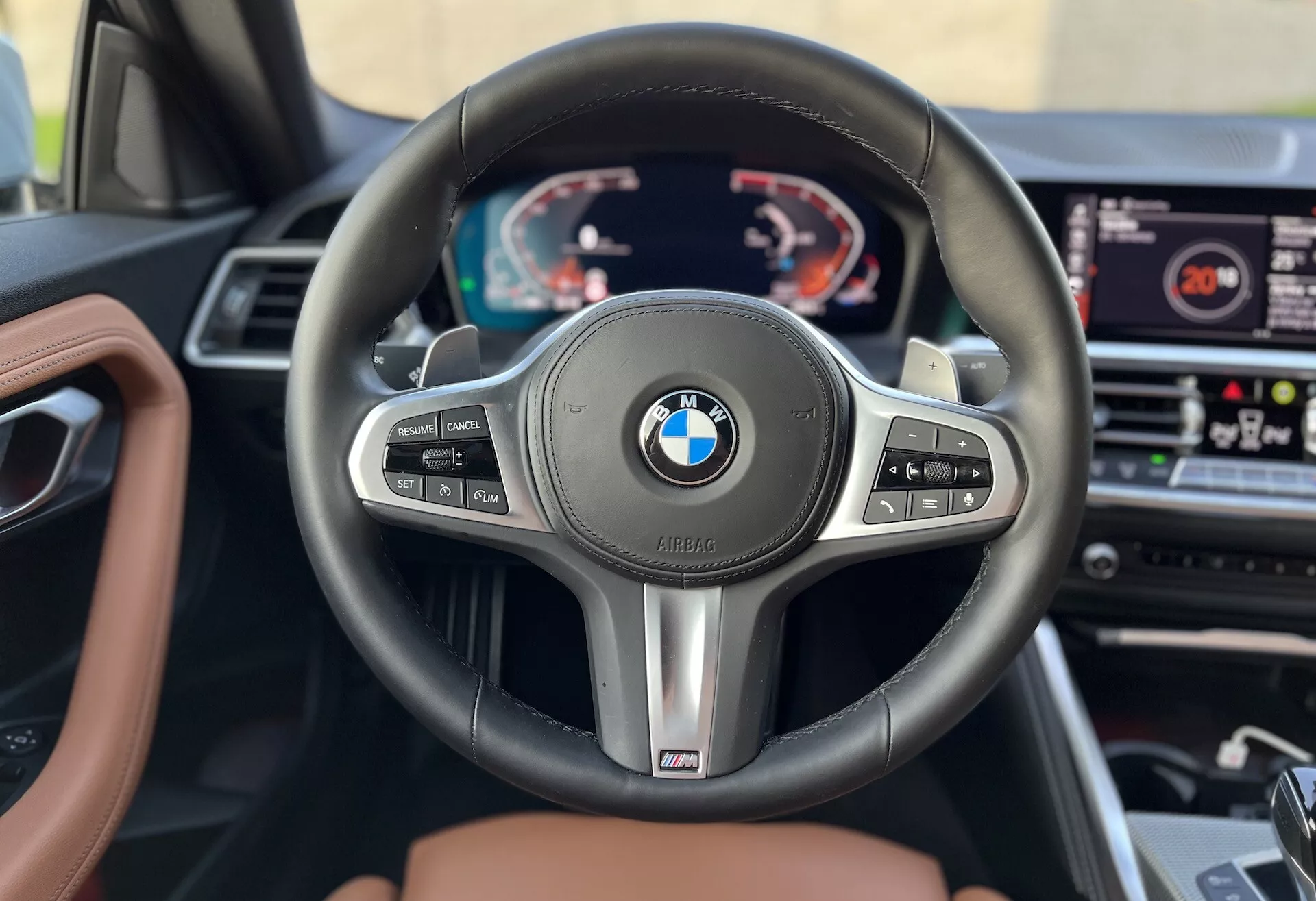 Belső kép egy 2022-es BMW 2-es sorozatú kupéról, amely a kormánykerékre és a műszerfalra összpontosít a vezetőülés perspektívájából. Jellemzője egy háromküllős BMW kormánykerék többfunkciós gombokkal a tempomat, az audio kezelés és a telekommunikáció számára, valamint váltófülekkel és egy 'M' teljesítmény jelvénnyel. Mögöttük egy digitális műszerfal található, amely egy fordulatszámmérőt, sebességmérőt és többfunkciós információkat jelenít meg. A műszerfal közepén látható az infotainment rendszer, és a belső díszítés bőrből és fából készült, jelezve egy prémium modellt.