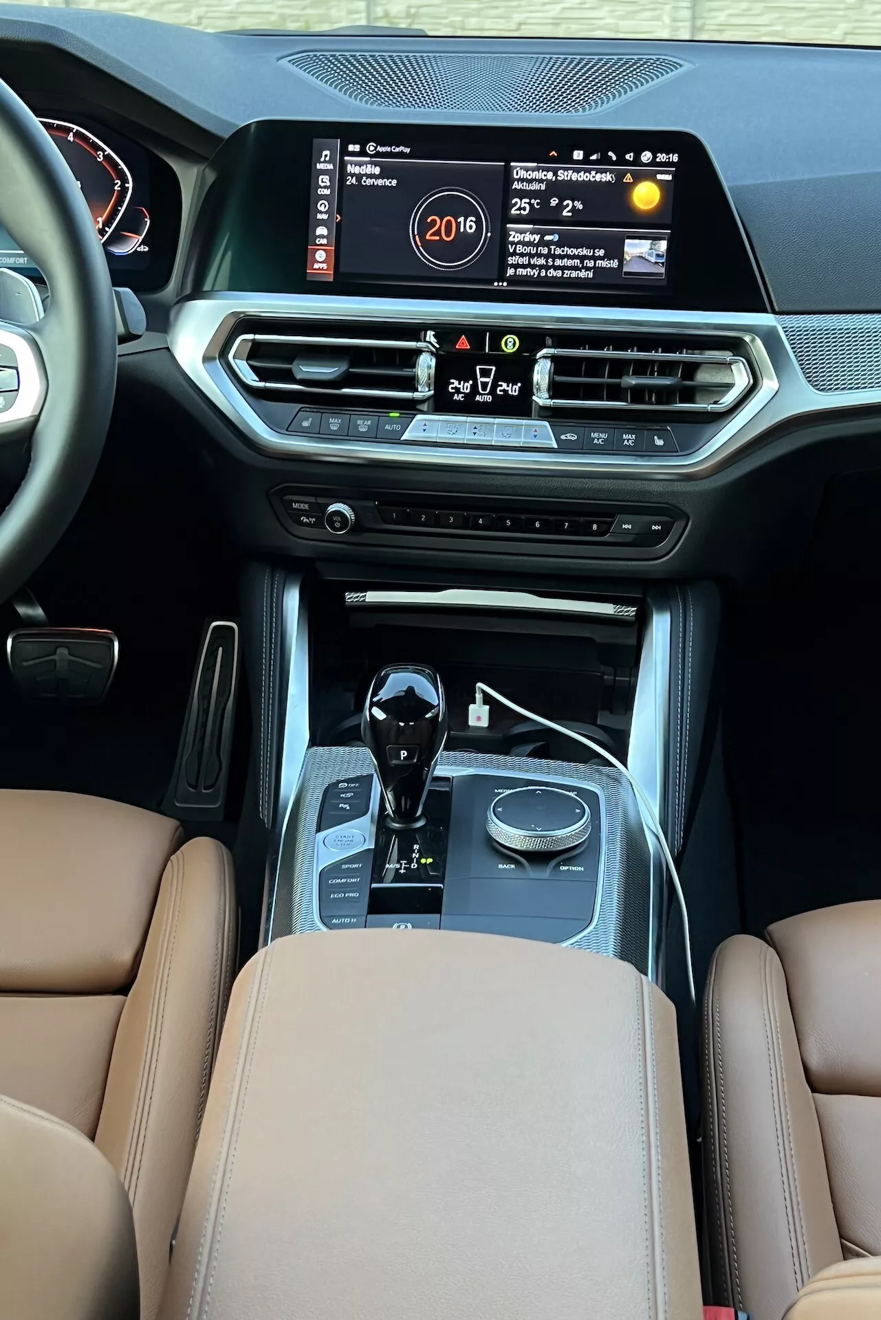 A 2022-es BMW 2-es sorozatú kupé belső nézete a hátsó utastéről, kiemelve a középkonzolt egy nagy infotainment képernyővel, amely az időt, a hőmérsékletet és a multimédiás rendszert mutatja. A jellemzők közé tartozik a légkondicionáló szellőzők, klímavezérlő gombok, indítógomb, automata sebességváltó bőrrel és krómozott díszítéssel, vezetési mód választók, barna bőrülések prémium kárpitozással és varrással, jelezve a luxust és a fejlett autóipari technológiát.