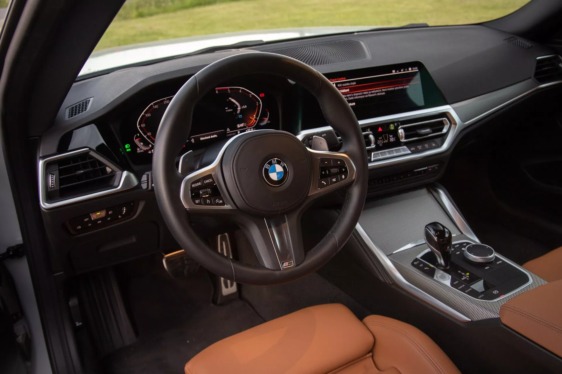 A 2022-es BMW 2-es széria kupé belső képe, amely a vezetői kabinra összpontosít. A magasabb perspektívából készített felvétel részletesen mutatja a multifunkciós kormánykereket BMW logóval, digitális műszerfalat, amely a sebességmérőt és a fordulatszámmérőt jeleníti meg, valamint a vezető felé orientált középkonzolt széles infotainment kijelzővel, klímavezérlő gombokkal, sebességváltókarral és iDrive vezérlőgombbal. Az ülések karamell vagy barnás színű bőrrel vannak kárpitozva, kihangsúlyozva a luxust és kényelmet.