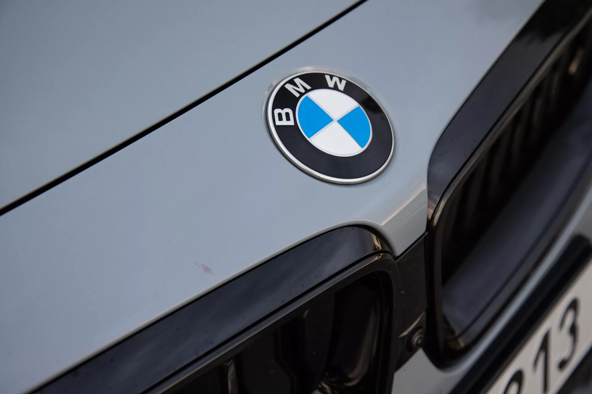 Közeli kép a 2022-es BMW 2-es széria Coupé elejéről, középen látható fekete, kék és fehér BMW emblémával a motorháztetőn. A kép a háttérben megjeleníti az ikonikus veserács felső szélét, az autó külső burkolata pedig fémszínű ezüst vagy világosszürke, ami visszaveri a környezeti fényt. A jelvényt éles fókuszban megörökítő fotó kiemeli a luxusmárkát és a jármű jól karbantartott felületét.