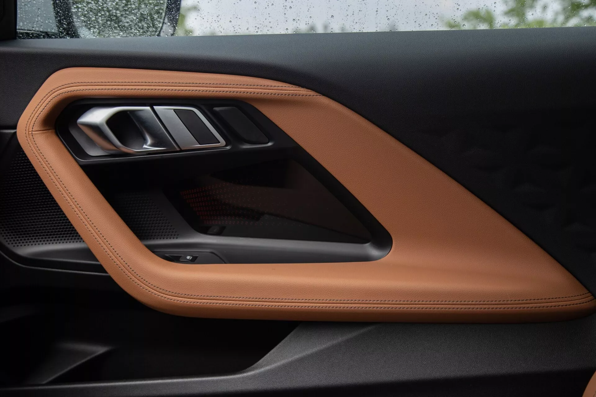 Közelkép a BMW 2 Series Coupe 2022 belső ajtópaneljéről, luxus varrott barna vagy konyak színű bőrrel, elegáns fém ajtókilinccsel és sportos karbonszálas díszítőelemmel, kiemelve a kiváló minőségű anyagokat és a kifinomult dizájnelemeket.