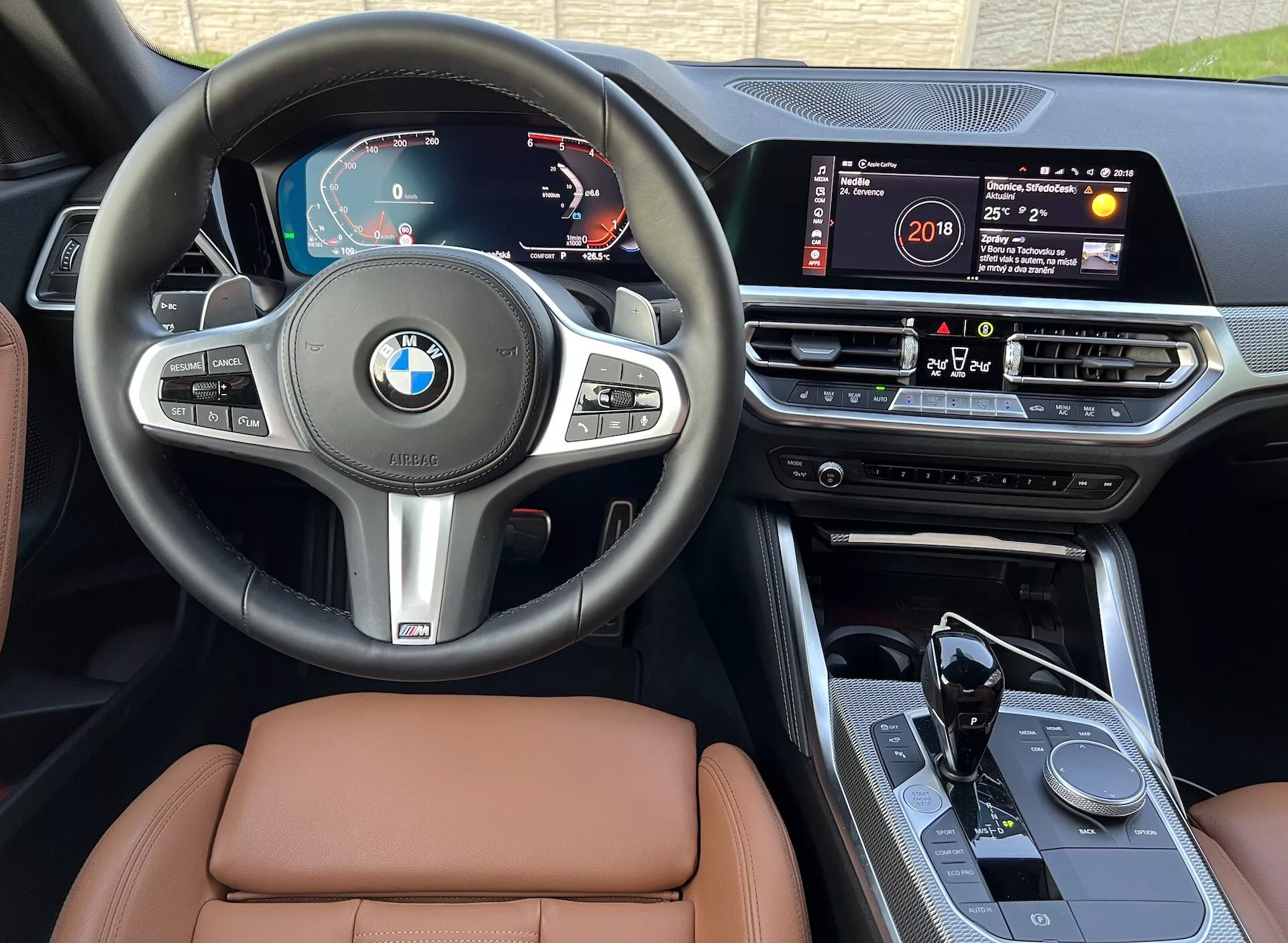 A 2022-es BMW 2-es sorozat kupé belső képét ábrázolja, amely a műszerfalat, a kormánykereket és a középkonzolt mutatja. Jellemzők közé tartozik a digitális műszercsoport, a multifunkciós kormánykerék, a nagyméretű érintőképernyős infotainment rendszer, a középkonzolon található forgó vezérlőgomb, az automata váltó kar sportos kialakítással és a magas színvonalú bőr ülések, mindezt a vezetőülés perspektívájából napfényben.