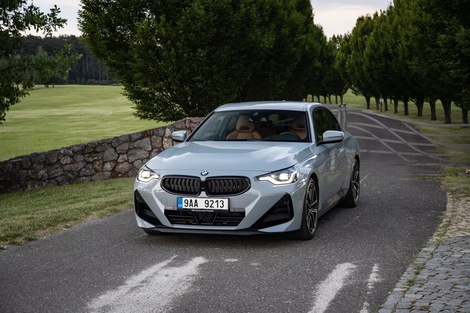 Részletes kép egy 2022-es BMW 2-es szériás Coupéról háromnegyedes első szögből, amely bemutatja az autó elejét és oldalát egy csendes aszfaltúton, amelyet gondozott fák és egy kőfal szegélyeznek. A jellegzetes BMW vesékrács és az elegáns LED fényszórók díszítik az elülső részt, míg a lökhárító légbeömlői sportos megjelenést kölcsönöznek neki. A motorháztető kontúros vonalai teljesítményt sugallnak, a jól megformált sárvédők erőt jeleznek, a többküllős könnyűfém keréktárcsák pedig a sportos kivitelre utalnak. Az oldalsó profil karaktervonala mozgást sugall, az oldalsó tükrök beépített irányjelzőkkel és egy sunroof villanása látható. A szélvédő szokásos dőlésszöge és a tiszta oldalablakok kiegyensúlyozott megjelenést kölcsönöznek az autónak. A lágy világítás, valószínűleg kora reggel vagy késő délután, finom árnyékokat és a forma és a szín tiszta bemutatását eredményezi.