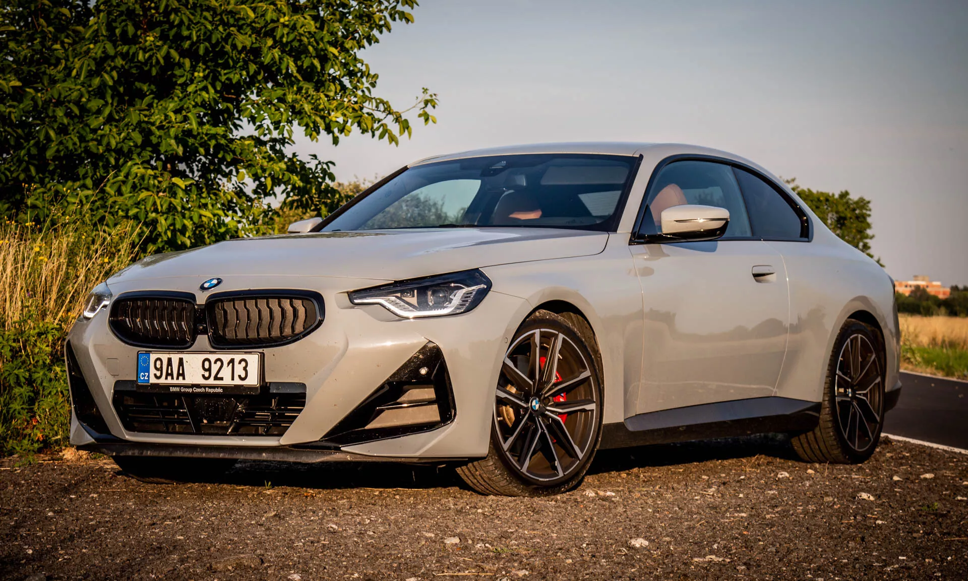 2022-es BMW 2-es sorozatú kupé képe. A modern luxusautó szögben parkol, kiemelve az autó bal oldali elejét aranyóra alatt. A felvétel a jellegzetes tervezési elemekre összpontosít, mint például a nagy vese alakú hűtőrács, szögletes LED fényszórók, sportos első lökhárító nagy légbeömlőkkel, valamint a többküllős fekete könnyűfém felnik magas teljesítményű féknyergekkel. Az aerodinamikus oldalsó tükörbe beépített irányjelzők találhatók. Az elegáns karosszéria és a prémium fényezés kiemelik az autó kompakt, sportos profilját és az alacsony tetővonalat.