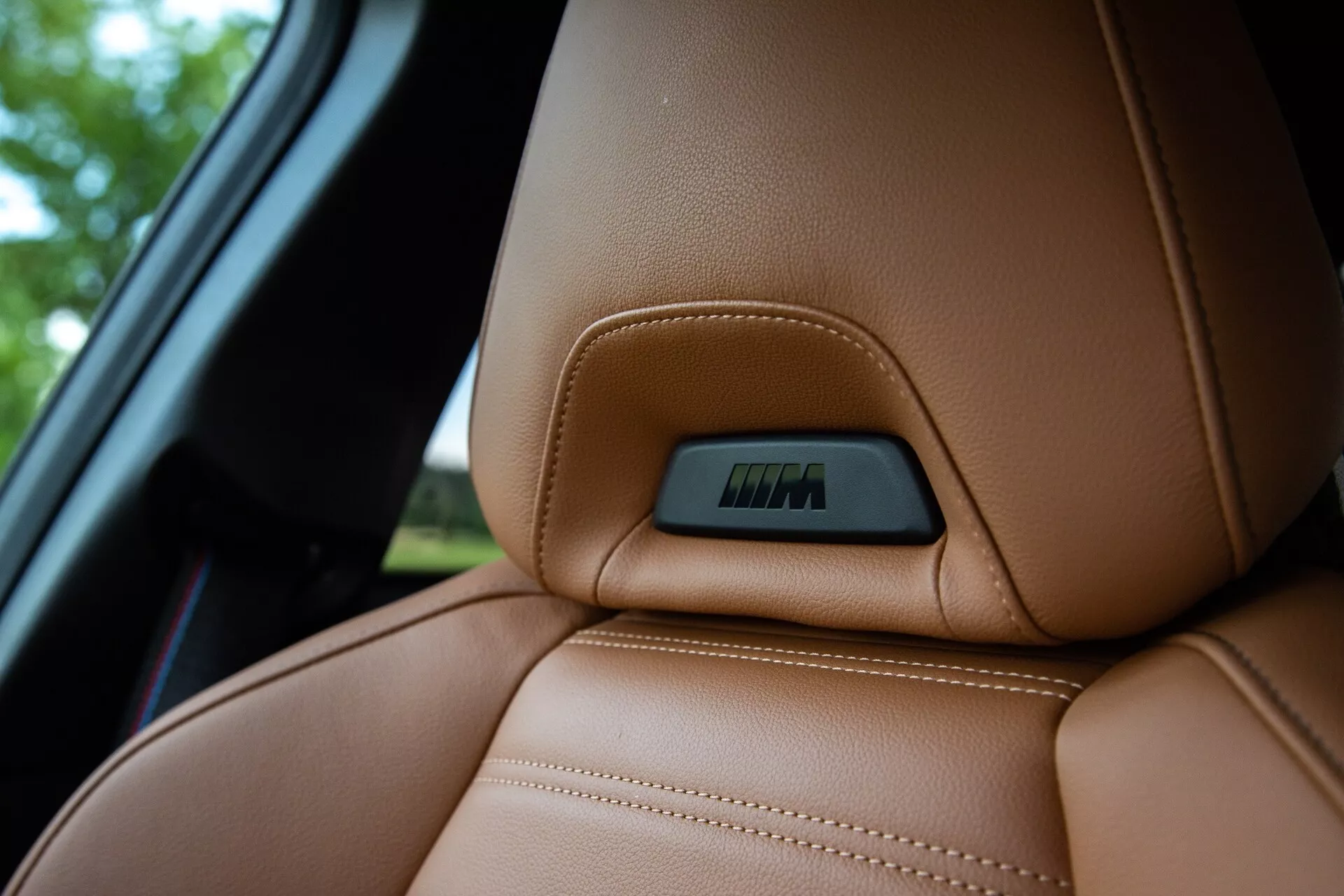 Közeli kép egy 2022-es BMW 2-es sorozatú kupé gazdag barna bőrhuzatának felső részéről, kiemelve a pontos, jól látható varrásokat és az ergonomikusan kialakított kontúrvonalat a kényelem és támasztás érdekében. Az ülés felső részébe beépített címke technológiai funkciókra, például elektronikus állításokra vagy fűtési funkciókra utal. A felvétel kissé alacsonyabb nézőpontból készült, kiemelve a bőr luxus textúráját, miközben a háttér elhomályosodik, amely magában foglal egy részt az autó ablakából.