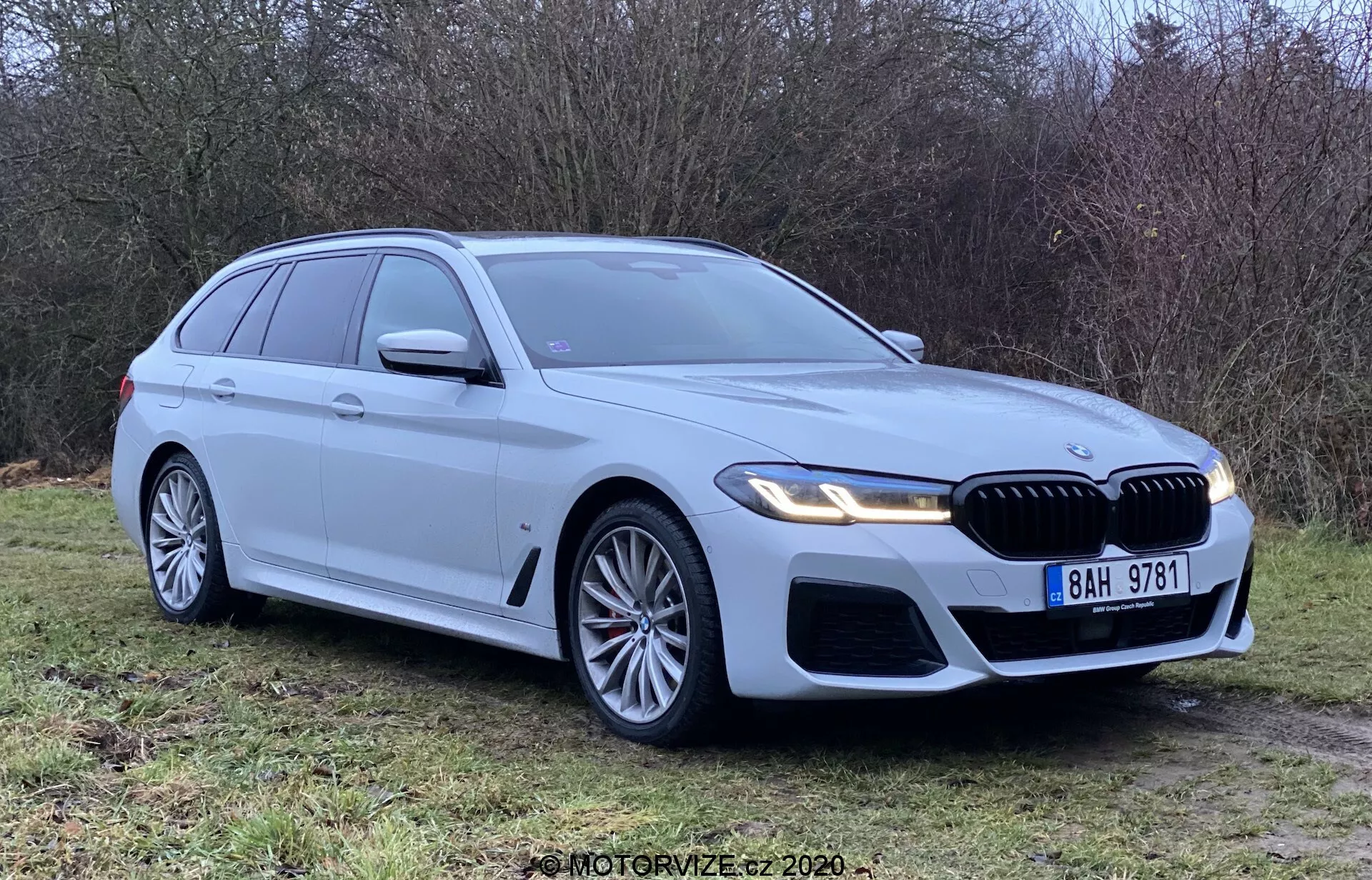 Ezüst BMW 5 Series Touring 2020 füvön parkol, háromnegyed elölnézetből nézve, megmutatva a hűtőrácsot, a fényszórókat és az aerodinamikus dizájnt. Jellemzői között LED fényszórók, nagyméretű könnyűfém kerekek, veserács és parkoló szenzorok. Tiszta és új, kiemelve a BMW jelvényt, integrált irányjelzőket, tetősíneket és cápauszony antennát.