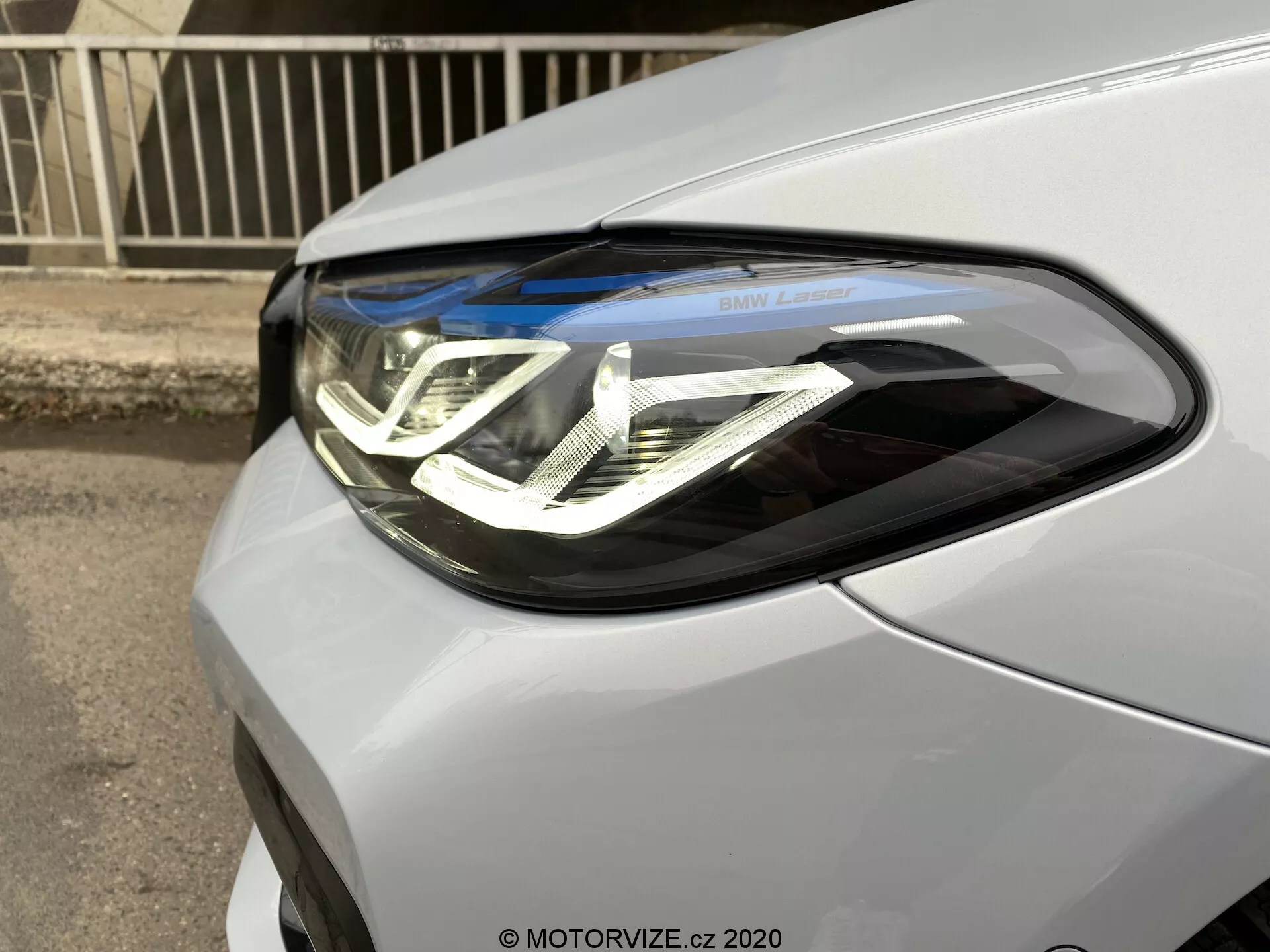 Közelkép a BMW 5-ös sorozat 2020 (BMW 520i Touring Steptronic) vezetőoldali fényszórójáról. A kép a járművet elölről, háromnegyedes szögből ábrázolja, kiemelve a bonyolult, dupla U alakú LED nappali menetfényt, a projektorlencséket és a „BMW Laser” feliratot. A fényszóró egység zökkenőmentesen integrálódik az autó metál vagy gyöngyházfényű fényezésébe.
